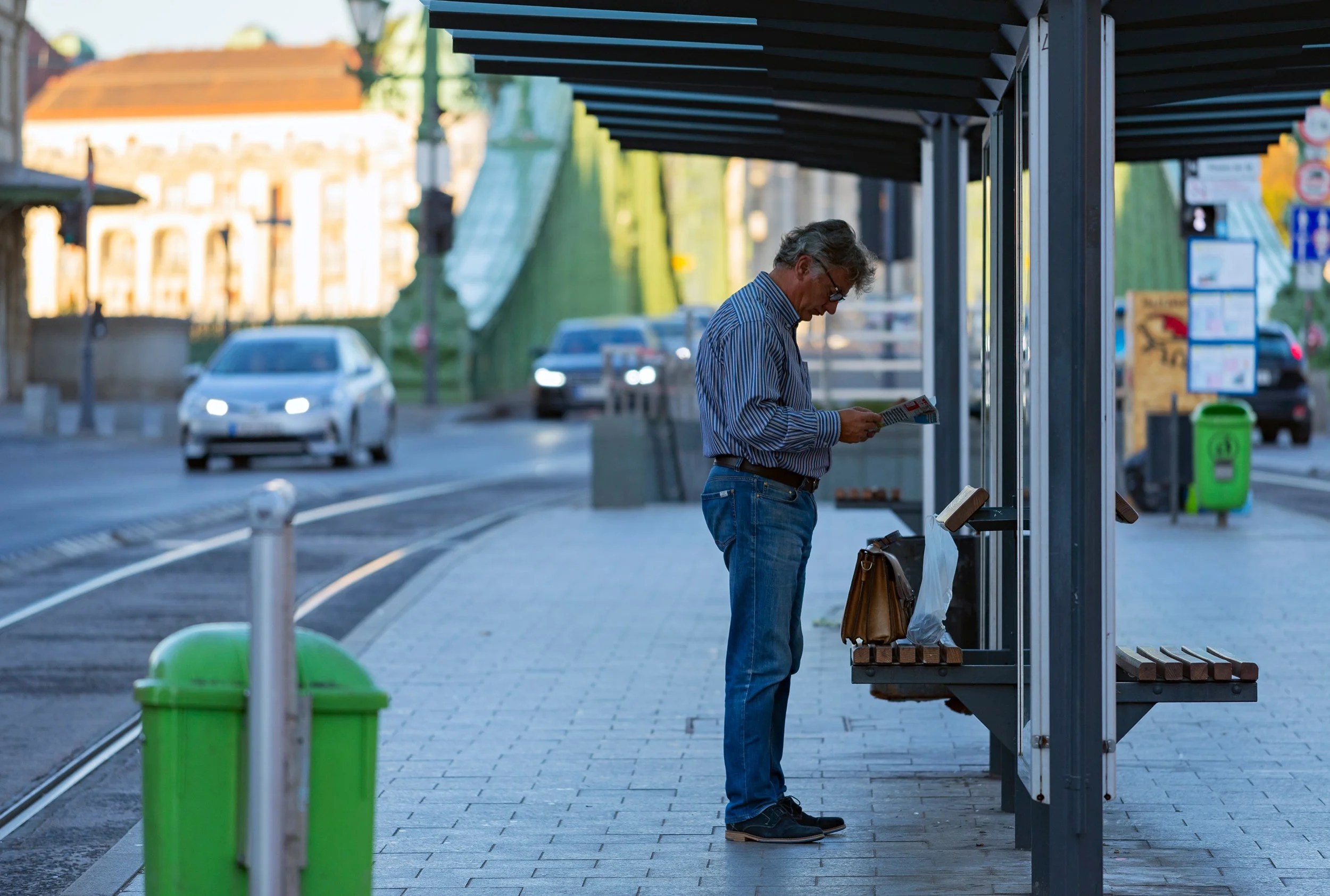 An elderly man with gray hair and glasses standing at a bus stop, reading a newspaper, with his brown bag on a wooden bench nearby. Cars are passing by in the background, and the scene is outdoors in a city setting.