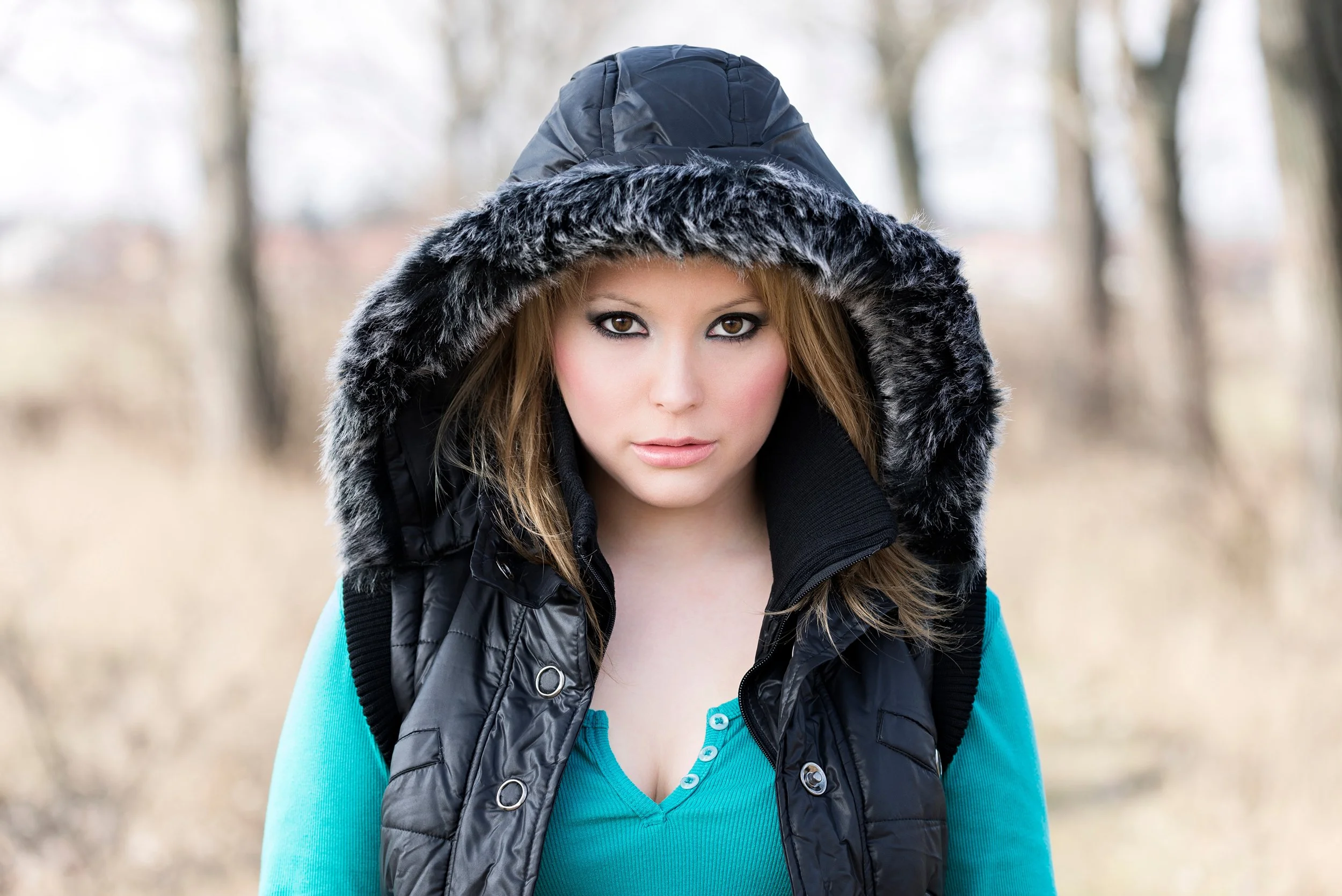 A woman with brown hair and brown eyes wearing a black jacket with a gray fur-lined hood and a blue long sleeve shirt, standing outdoors in a blurred wooded background.