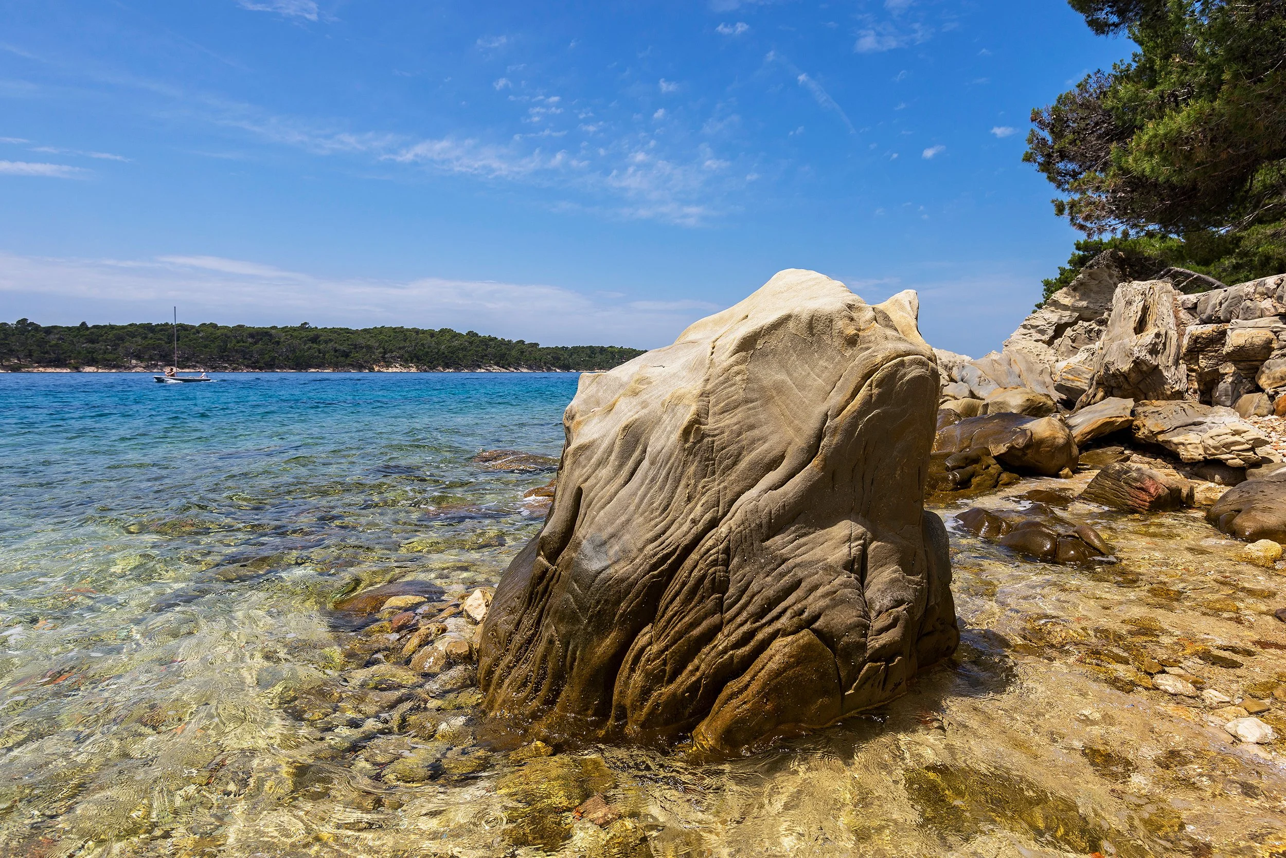 Peter Nemeth photographer. A rocky shoreline with large stones and clear shallow water, a sailboat floating on the blue sea, a green forested island in the background, and a bright blue sky with a few scattered clouds.