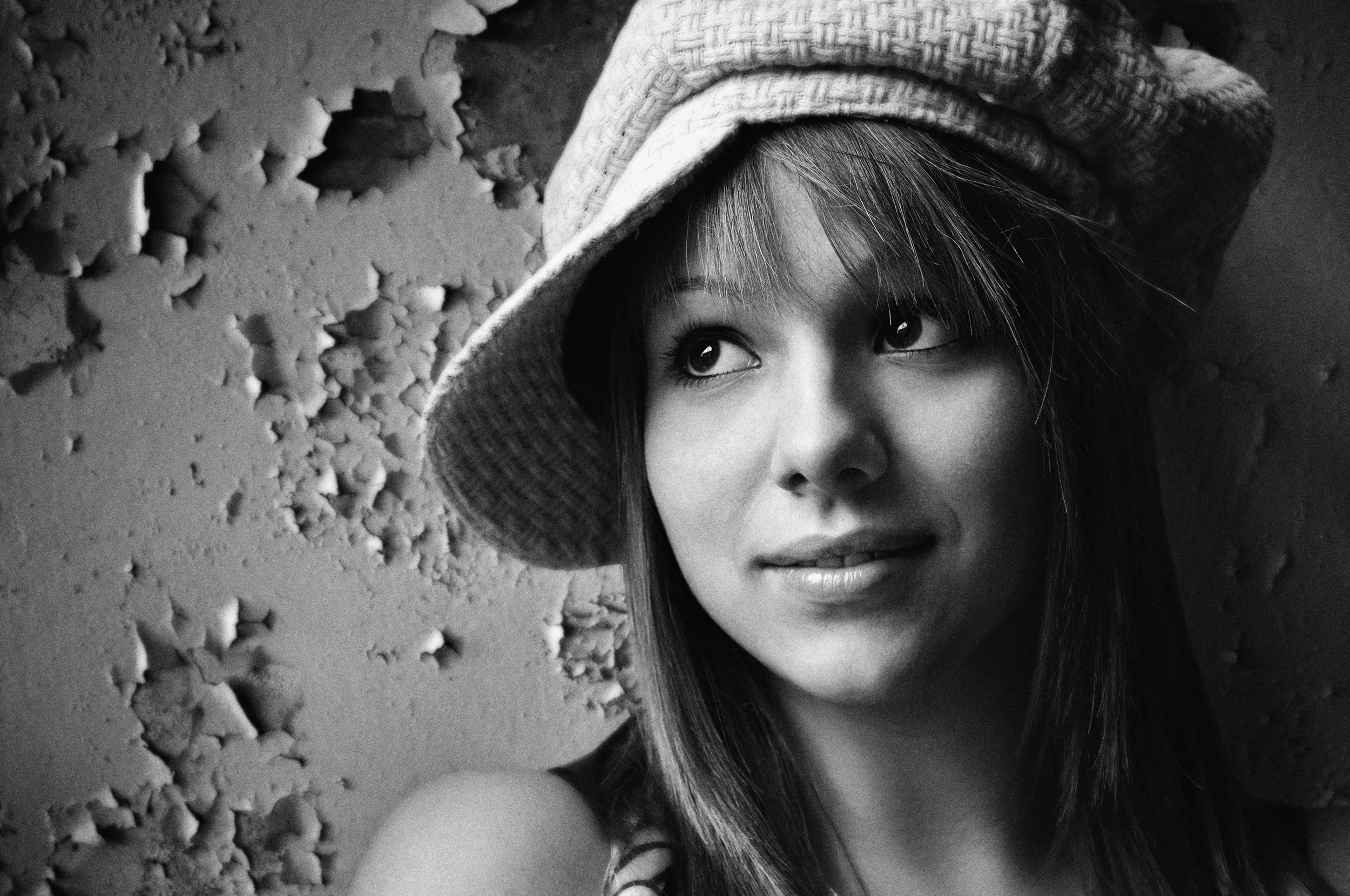 A young woman with long hair wearing a knit hat, looking slightly upward, with a textured wall in the background.