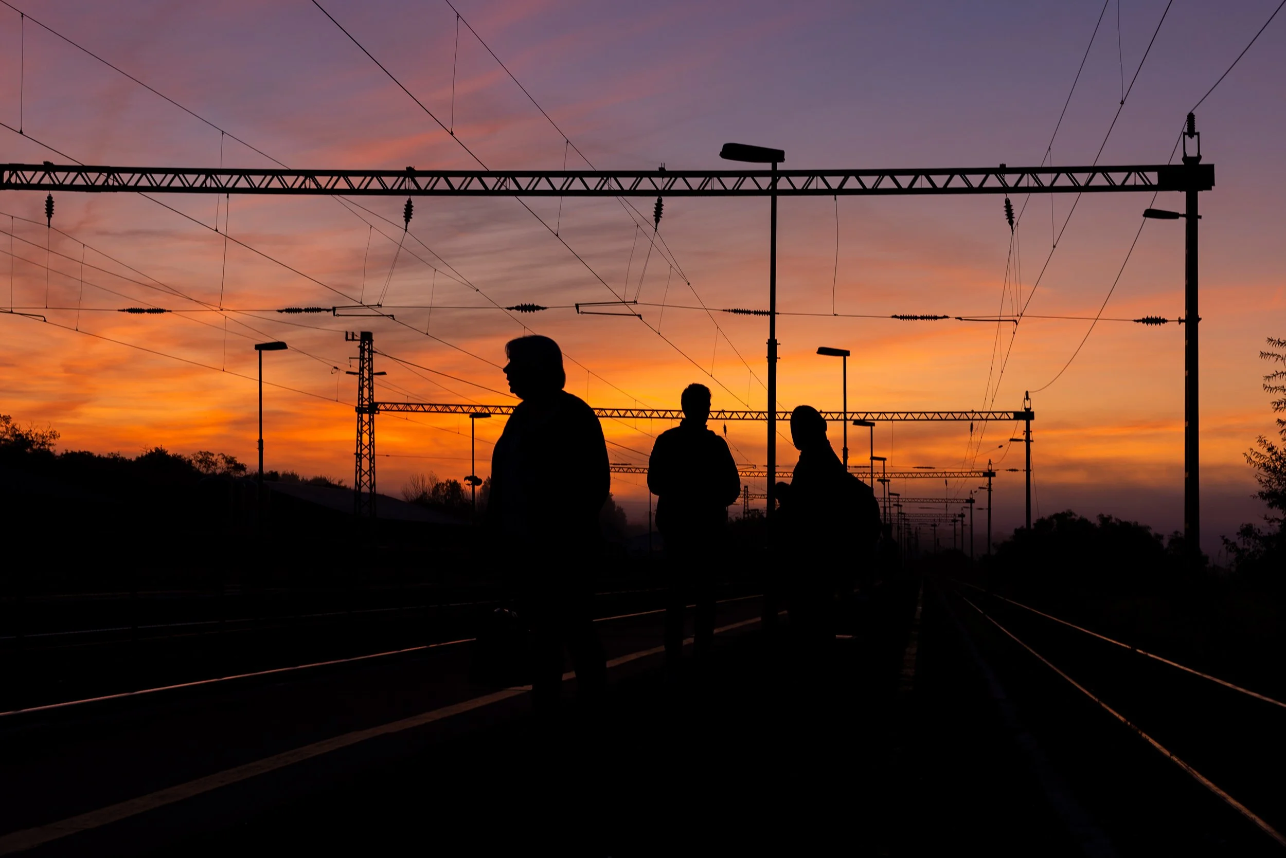Silhouettes of three people standing on railway tracks during sunset with electrical wires overhead and streetlights.