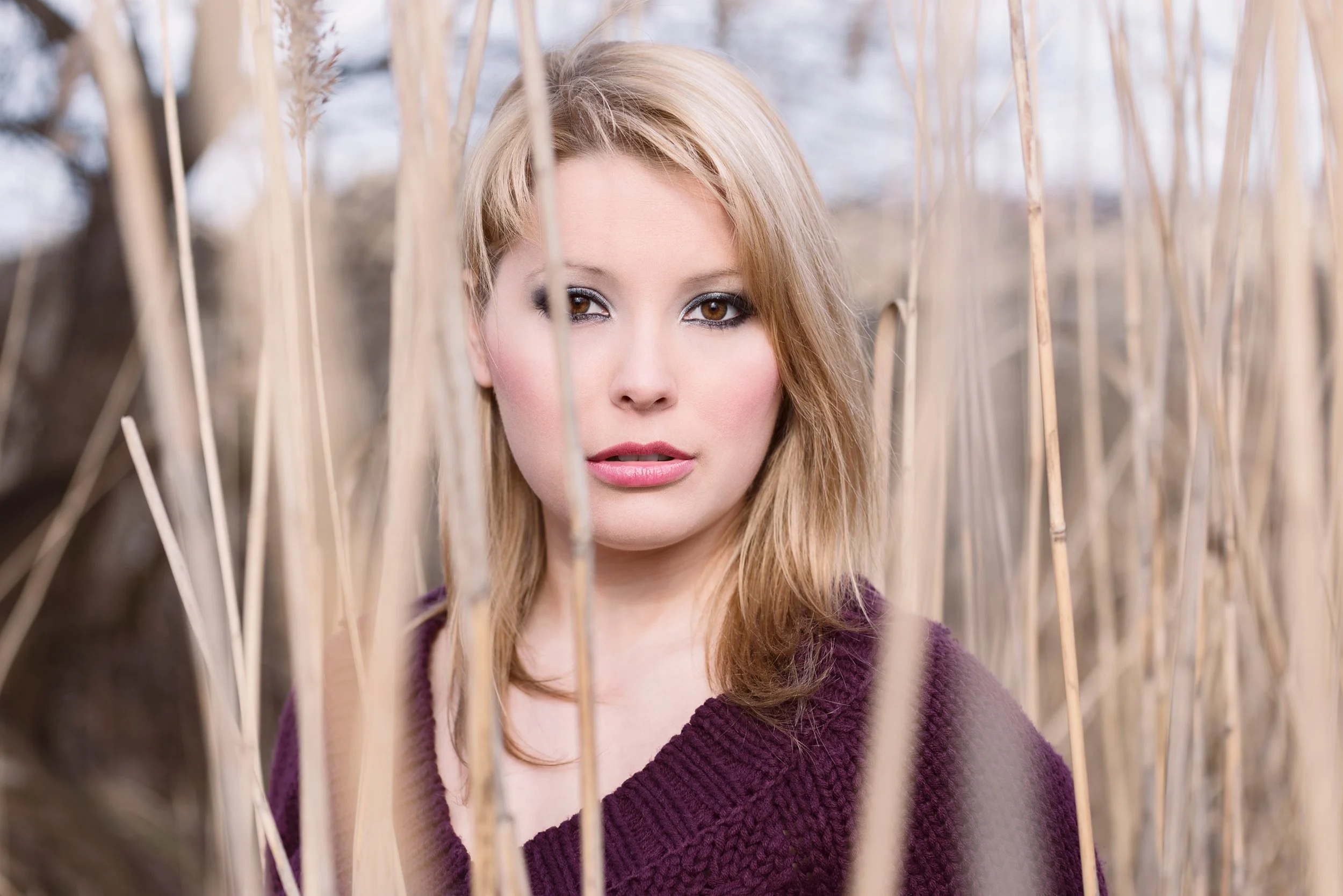 A young woman with blonde hair and makeup looking through tall, dry grass outdoors during fall or winter with trees in the background.