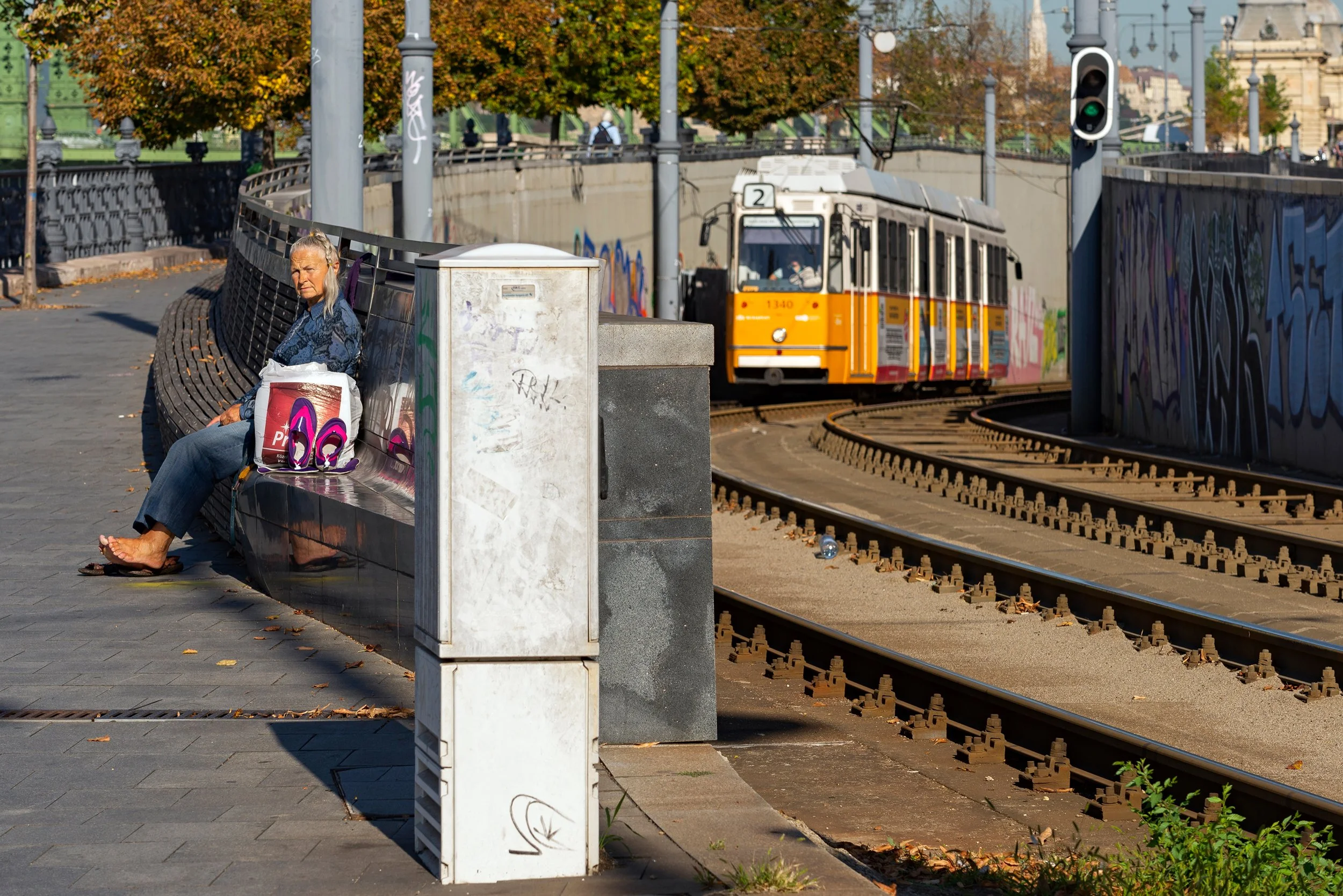 A woman sitting on a park bench with shopping bags and purple sandals, near train tracks with an orange train approaching, graffiti on wall, trees with fall foliage, light poles, and traffic signal.