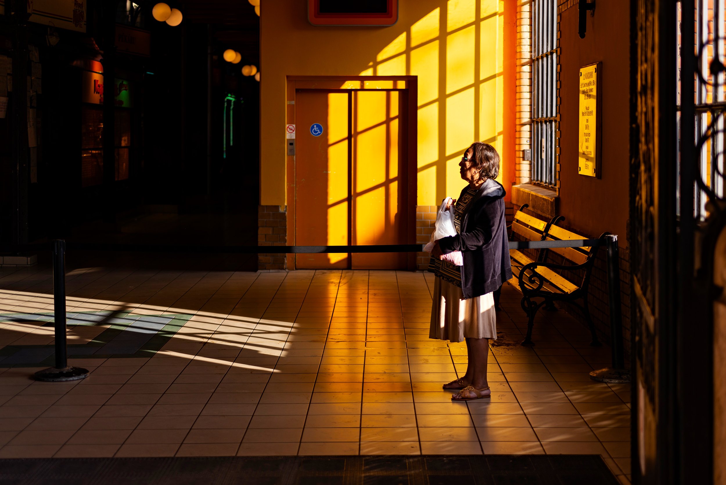 A woman standing alone inside a train station or terminal, illuminated by warm sunlight casting shadows on a yellow wall. She is holding a shopping bag and wearing glasses, with a vacant expression. There are benches, a sign, and barred windows nearb