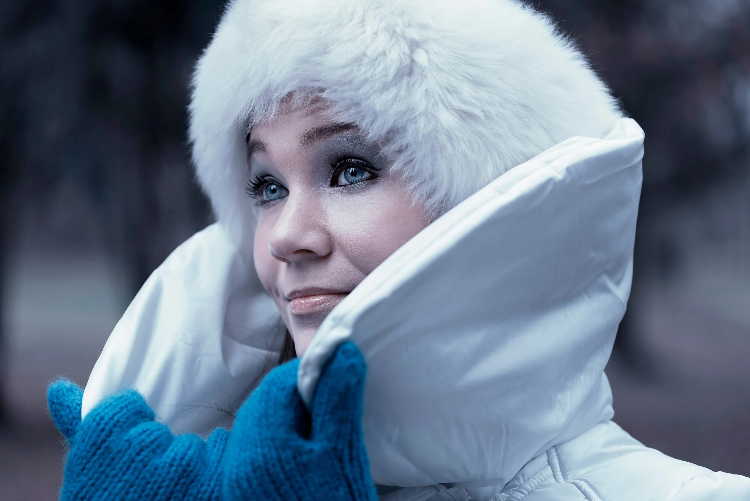 A woman dressed in winter clothing, wearing a white fur hat and coat, with blue gloves, outdoors in a cold environment.