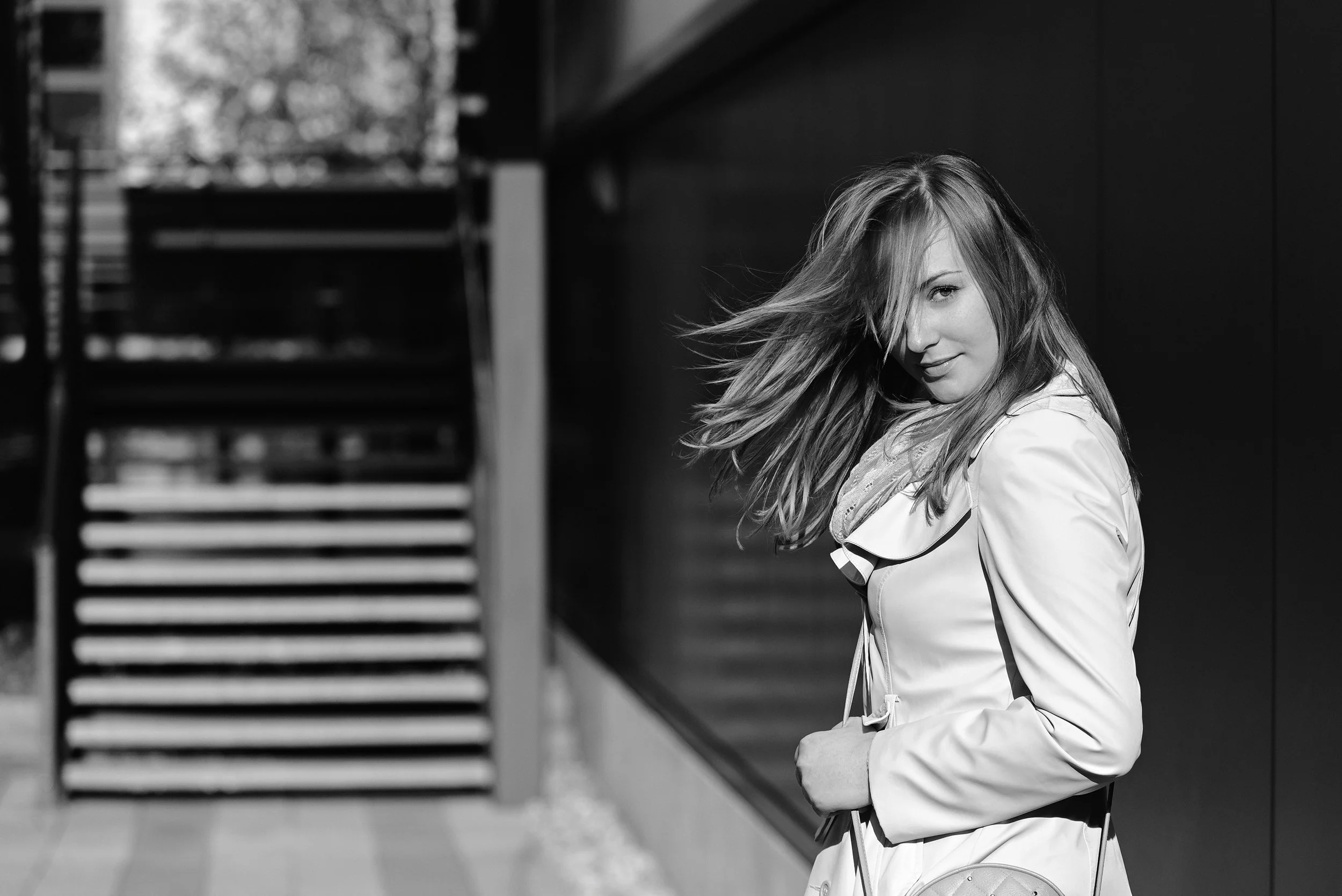 A young woman with long hair sitting on a bench outside, with her hair slightly covering her face, wearing a light-colored jacket, in black and white photograph.