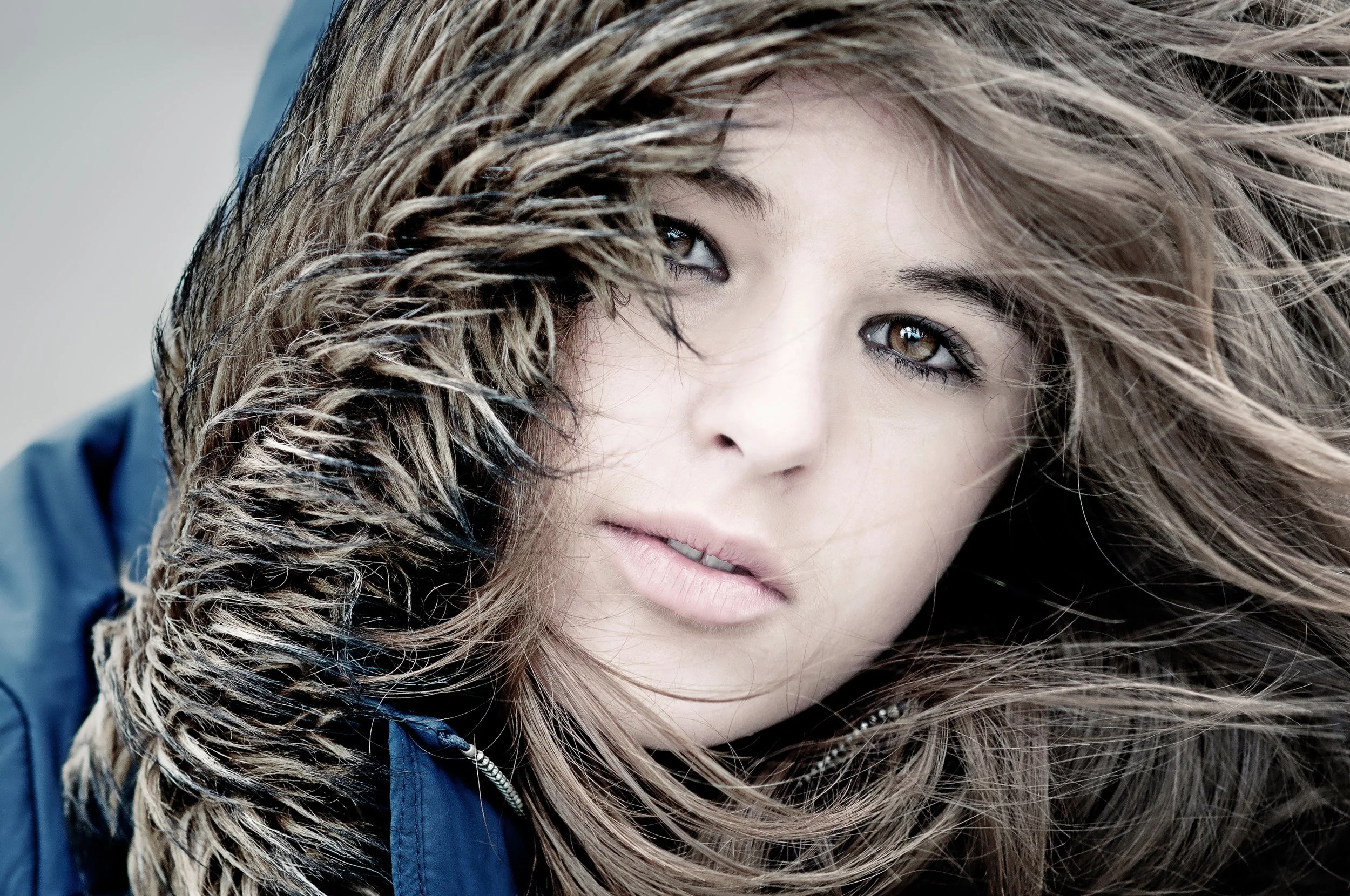 A close-up of a woman with light brown hair and hazel eyes, wearing a blue jacket with a fur-lined hood.