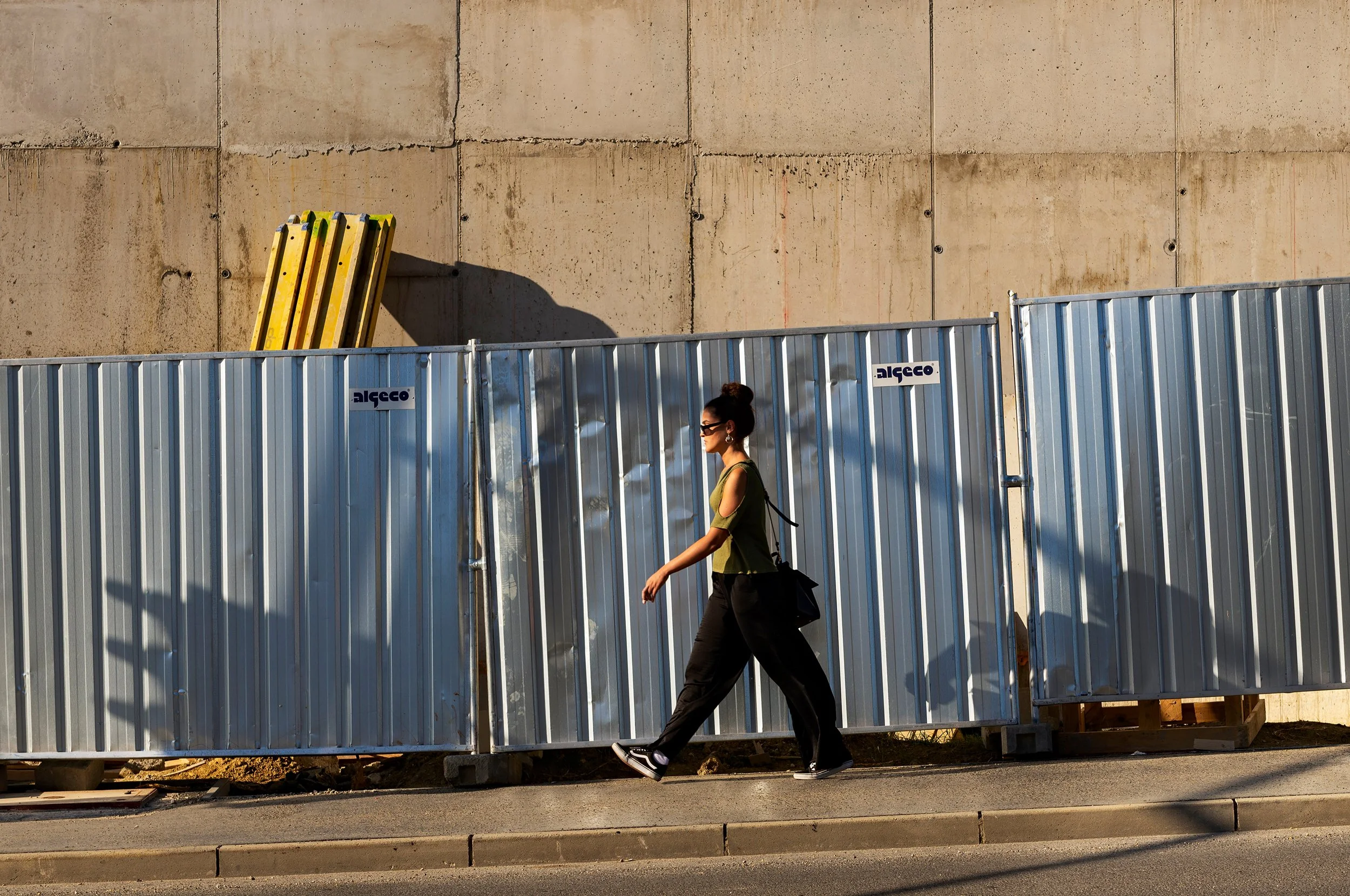 A woman walking along a construction site sidewalk in front of a tall, beige concrete wall, with a metal construction barrier labeled 'algeco' in front of her.