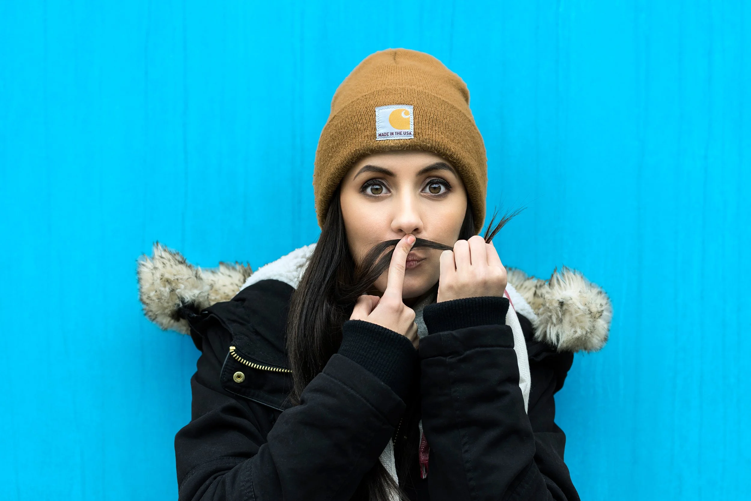 Young woman with brown eyes wearing a tan Carhartt beanie, a black winter jacket with a fur-lined hood, holding a lock of her long dark hair against a bright blue background.