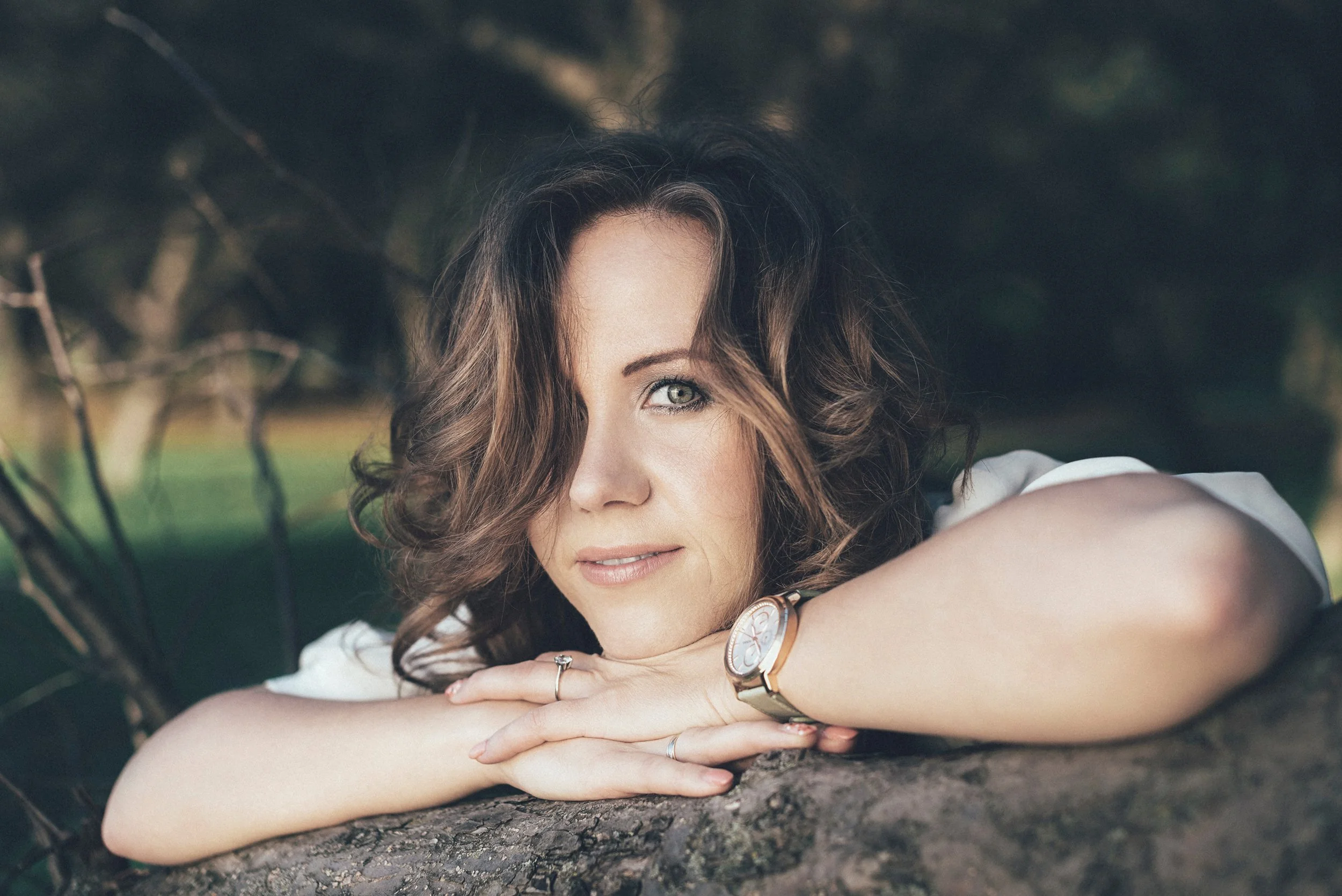Close-up of a woman with wavy brown hair, lying on her arms with her head resting on her hands, outdoors beneath trees.