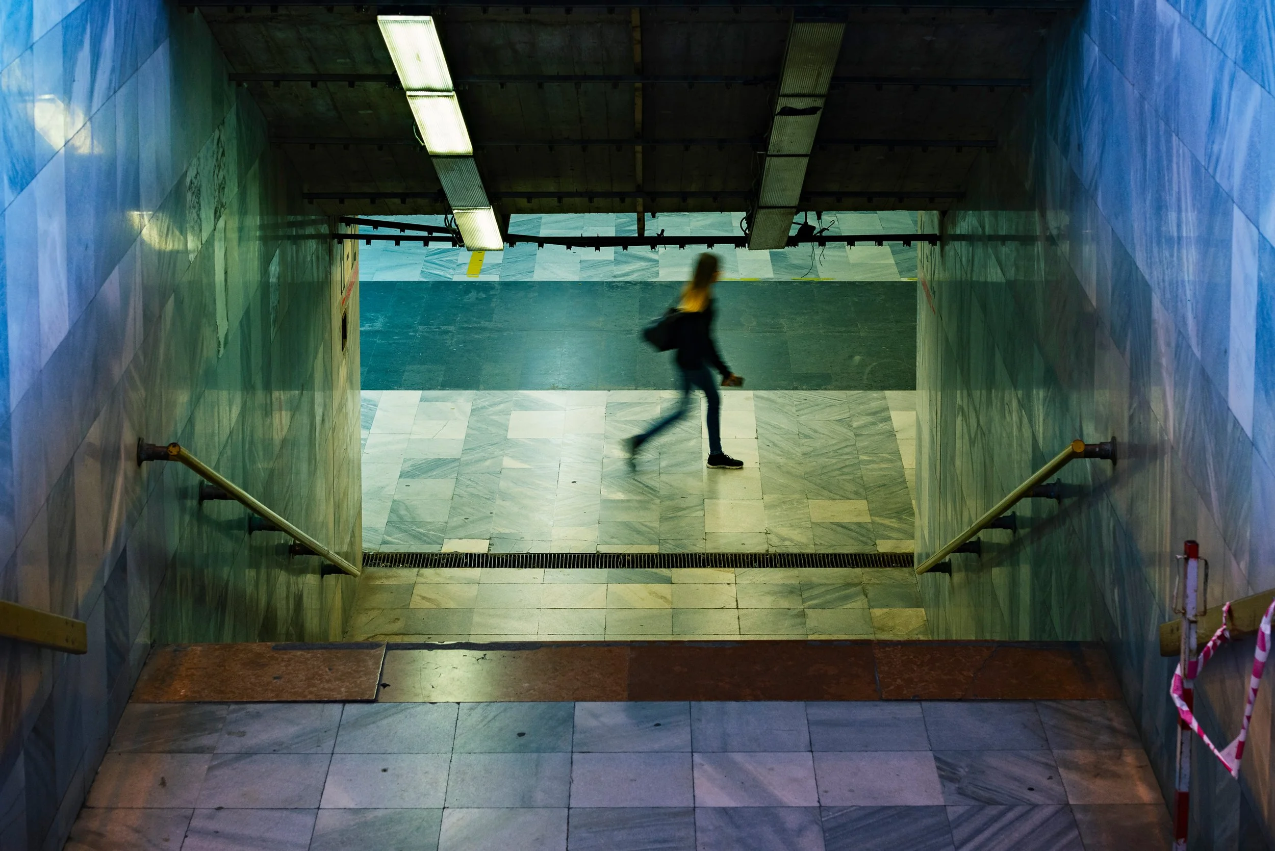A woman walking down a set of stairs in a corridor with marble tiles and green-tinted walls. The corridor is lit by fluorescent lights on the ceiling.