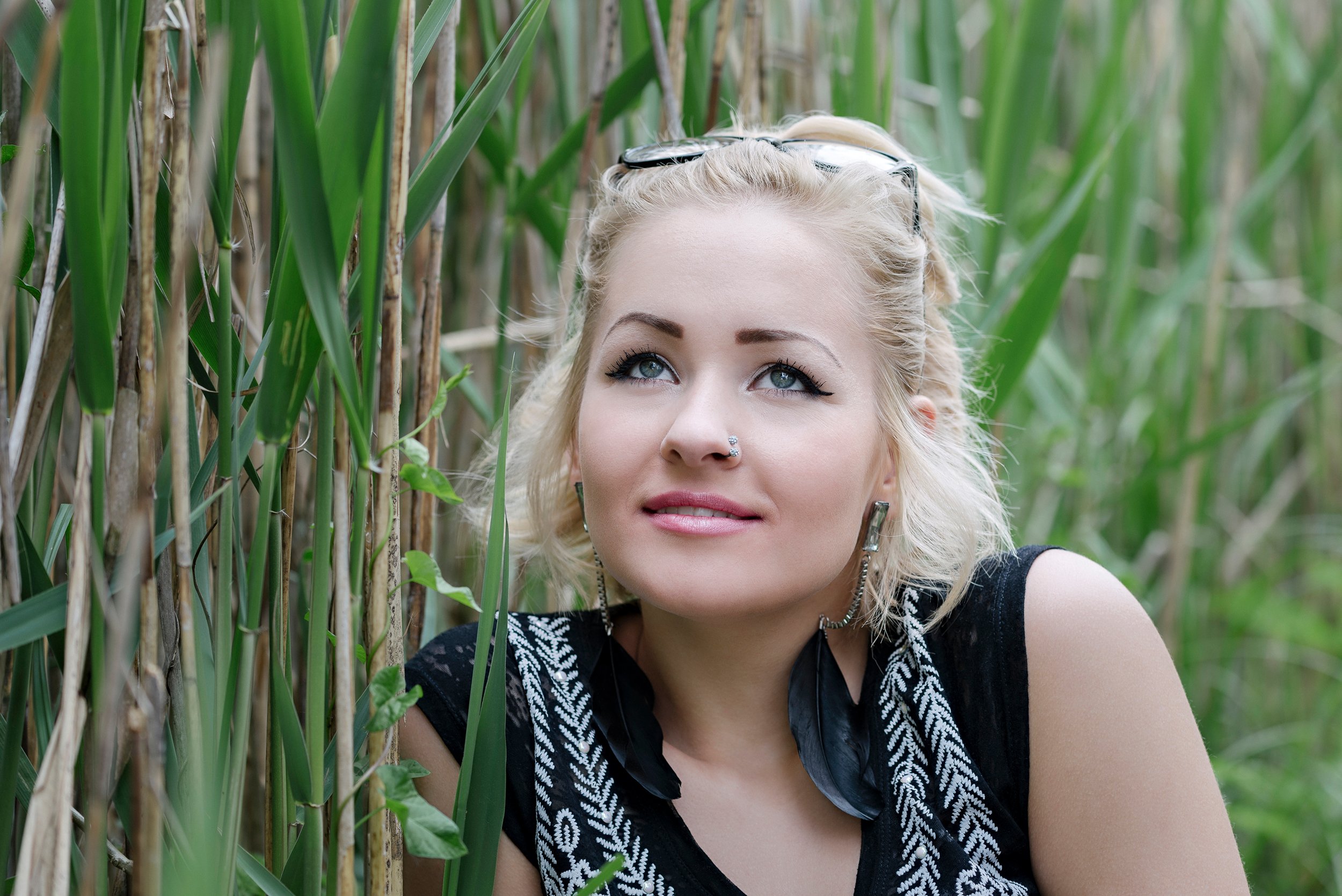 A woman with blonde hair, blue eyes, and a nose ring, wearing glasses on her head and a black and white patterned shirt, looking up while surrounded by tall green grass or crops in a field.