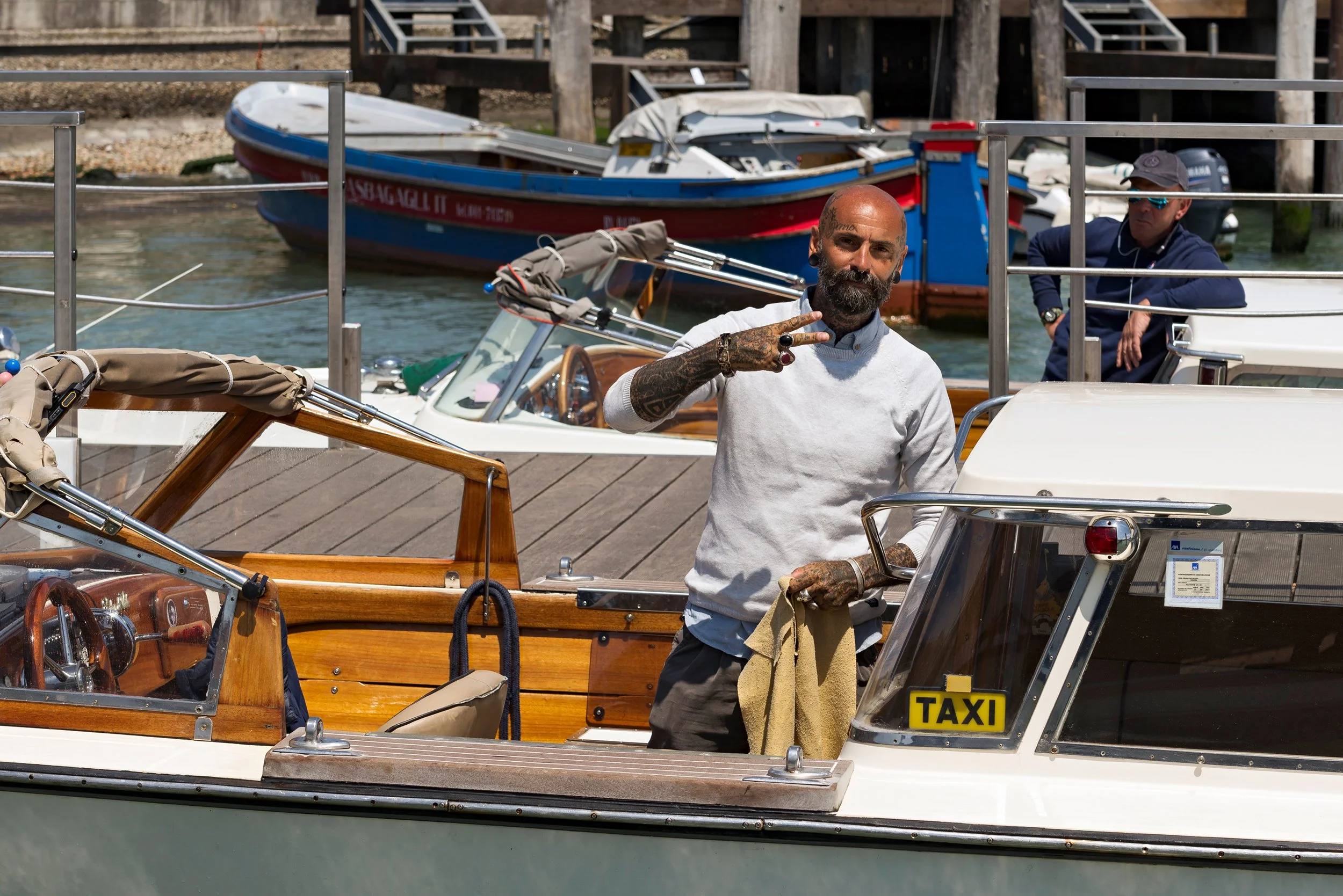 Man with tattoos and a beard making a peace sign on a boat docked next to other boats, including a small boat and a taxi boat, with boats and wooden pier structures in the background.