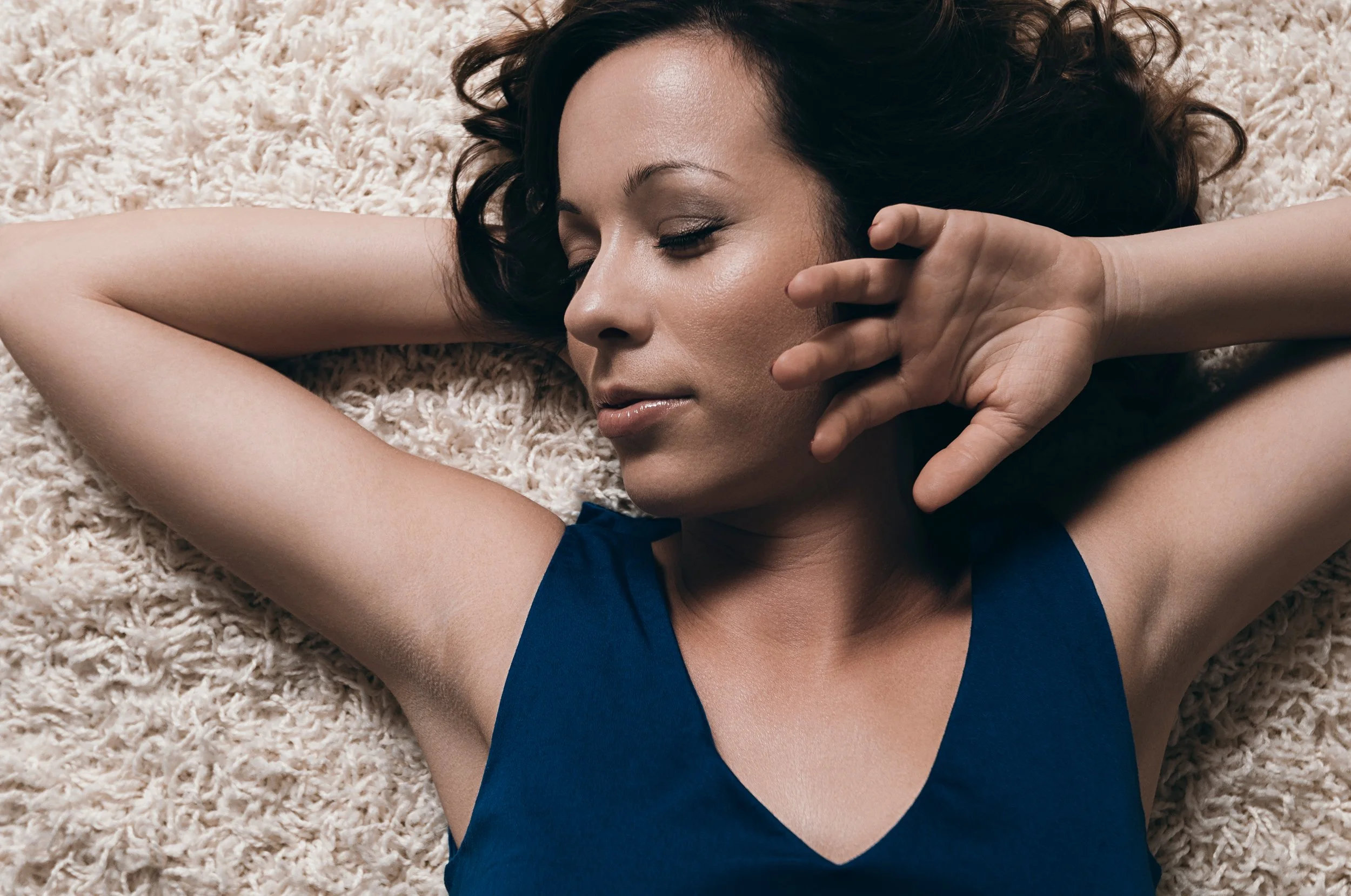 A woman with dark curly hair lying on a plush carpet with her eyes closed, facing slightly to the side, wearing a blue top and resting her head on her arm with her hand near her face.