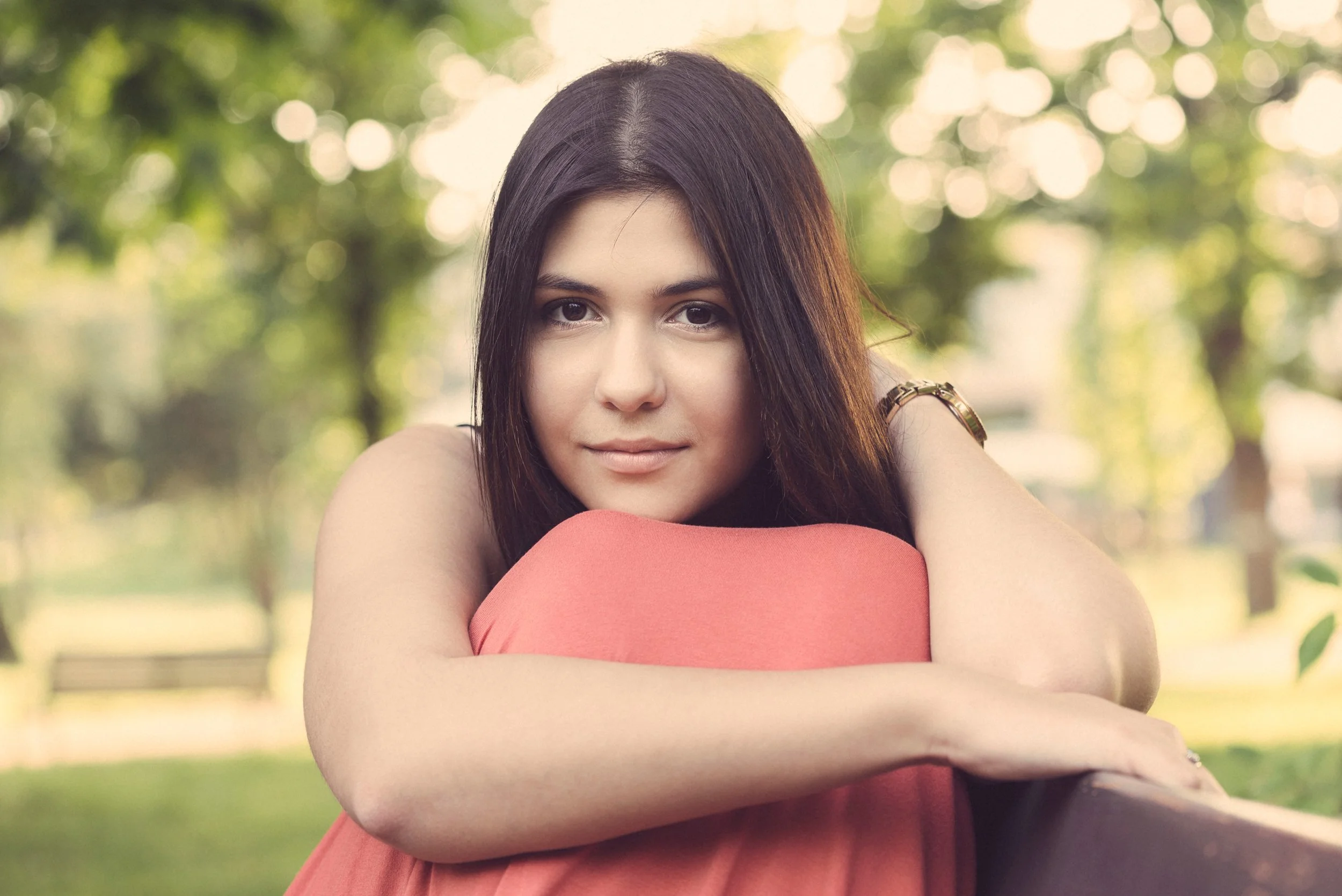 A young woman with long dark hair, resting her chin on her arms, smiling gently in an outdoor park with blurred green foliage in the background.
