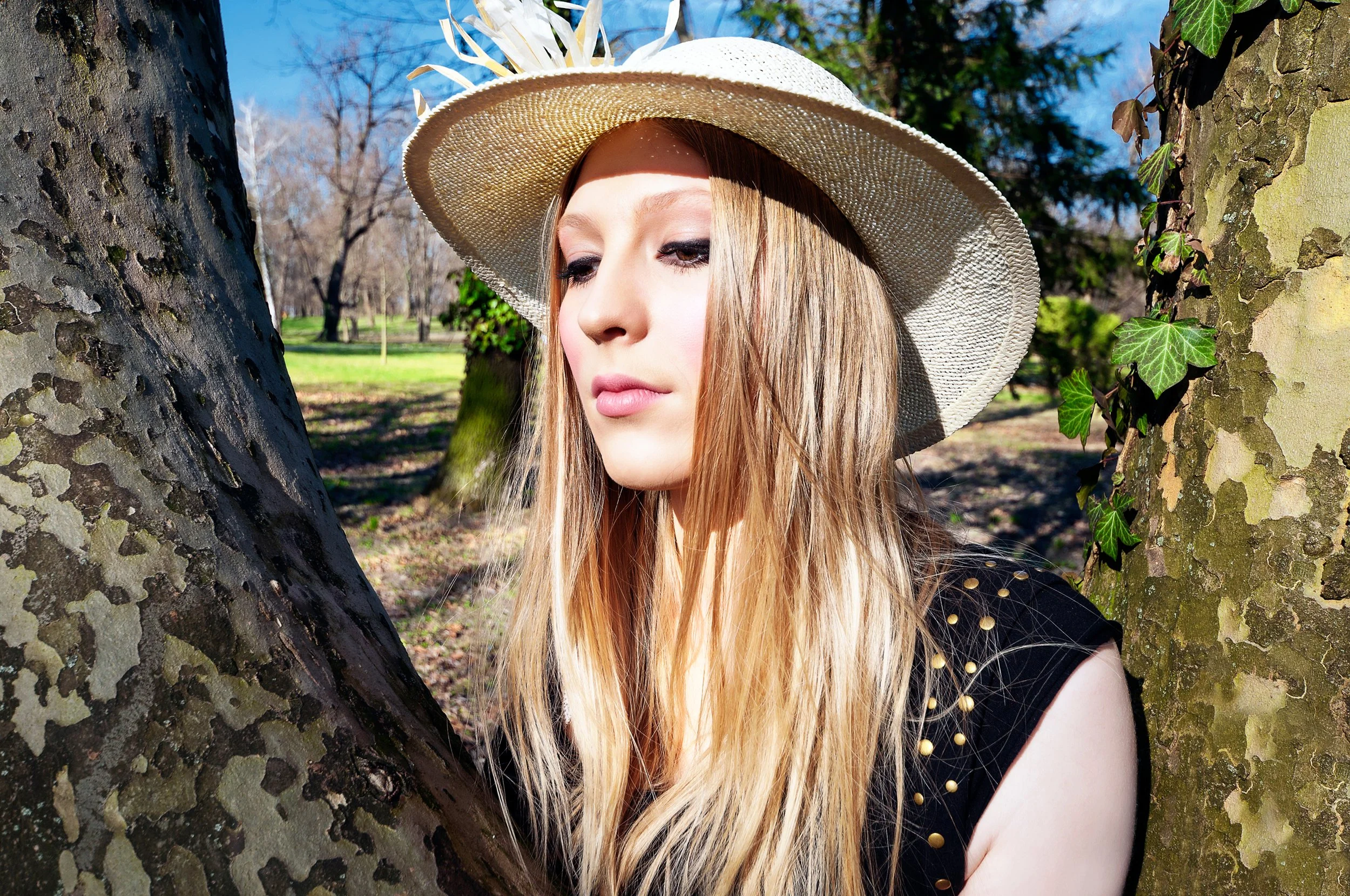 A young woman with long blonde hair wearing a wide-brimmed straw hat with decorations, standing outdoors between two tree trunks, with a park and blue sky in the background.