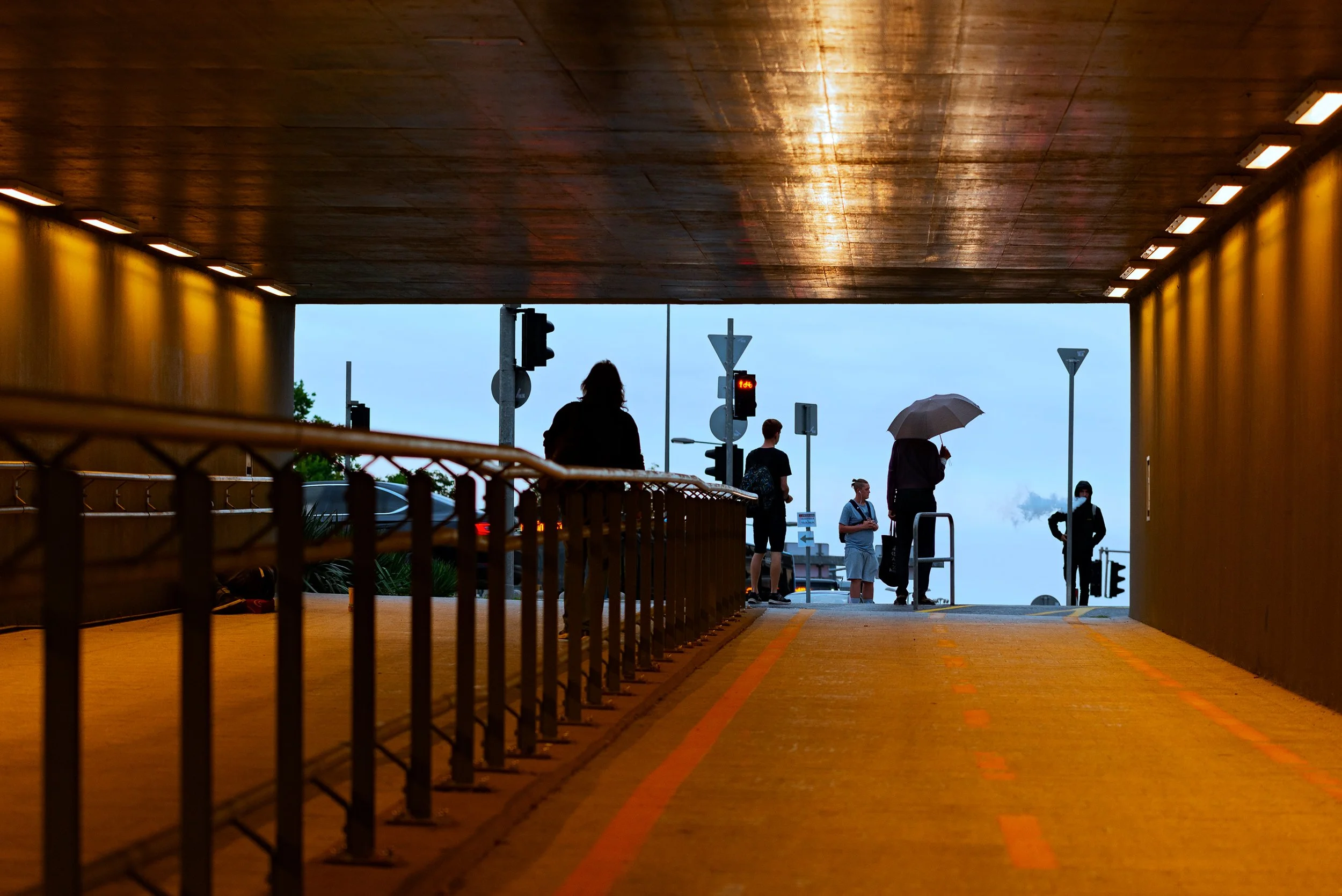 People standing and walking under a bridge with wooden ceiling and bright lighting, some using umbrellas.