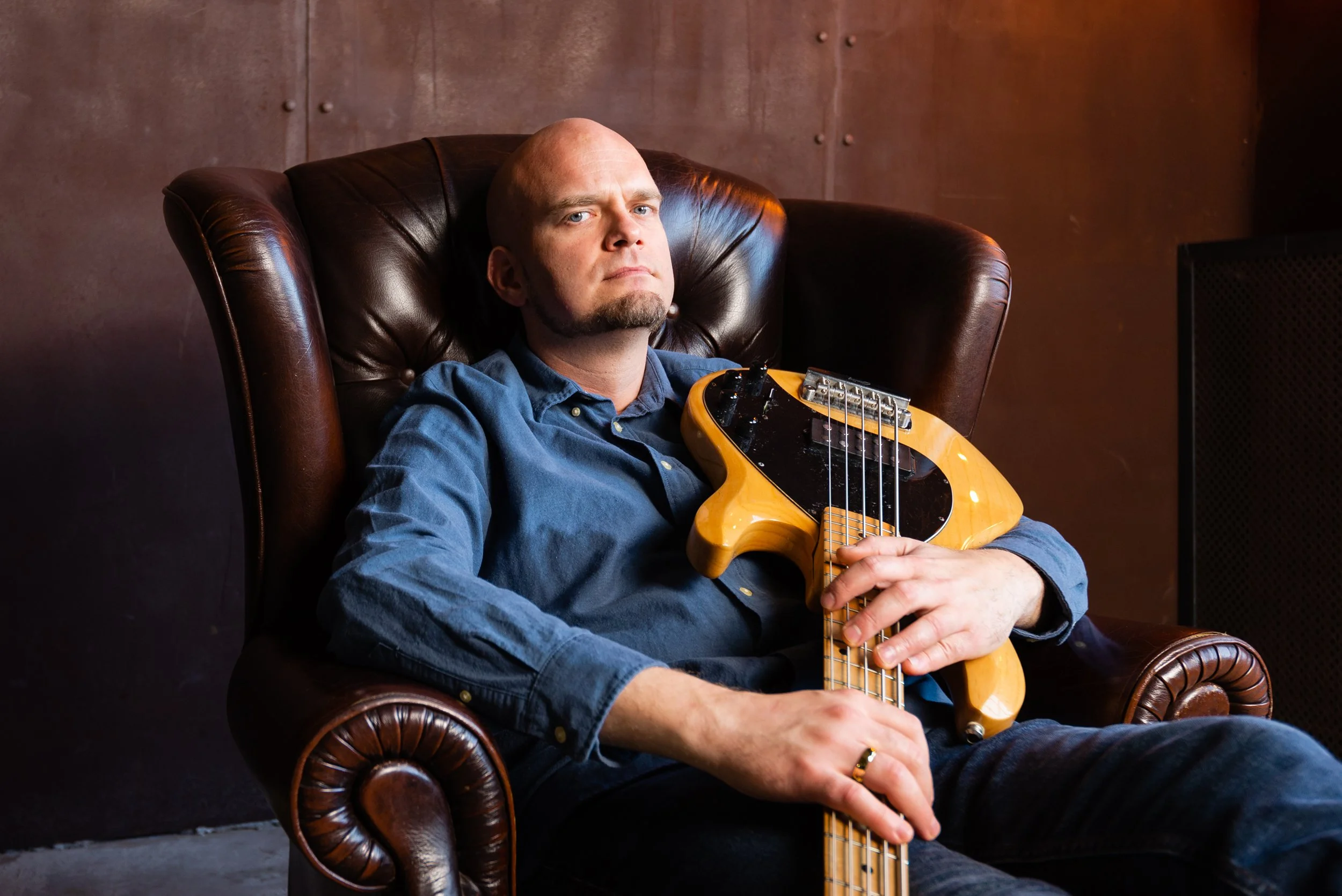 A man with a shaved head and beard sitting in a leather armchair, holding an electric bass guitar, in a room with brown walls.