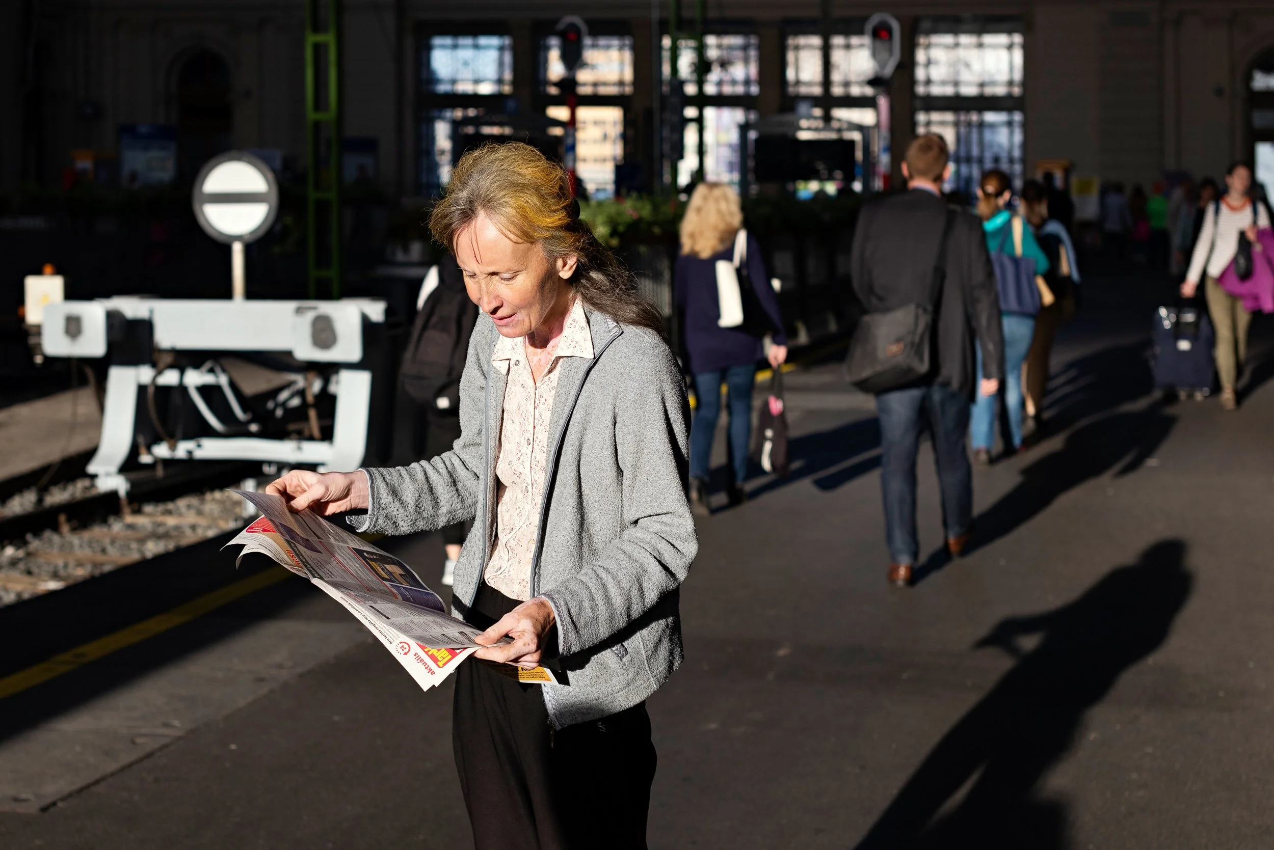 A woman in a gray jacket looking at a newspaper while standing on a train station platform. Other travelers with luggage are visible in the background, with some walking and some standing. The station has large windows and a train track.