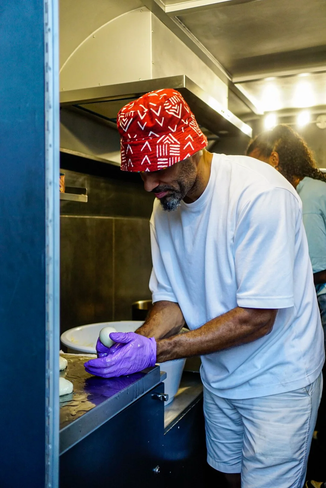 Beni Tonka wearing a red bucket hat, white t-shirt, and purple gloves is preparing dough in his foodtruck.