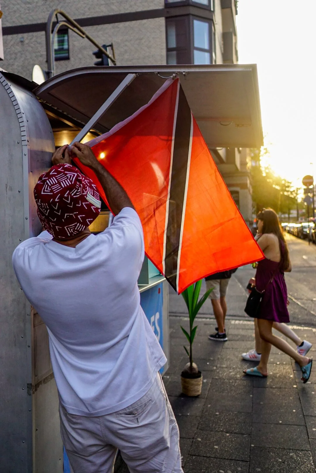 A man wearing a red patterned headscarf and white shirt is securing a large orange umbrella outside a building during sunset. In the background, a woman in a purple dress walks past, with other pedestrians and city buildings visible.