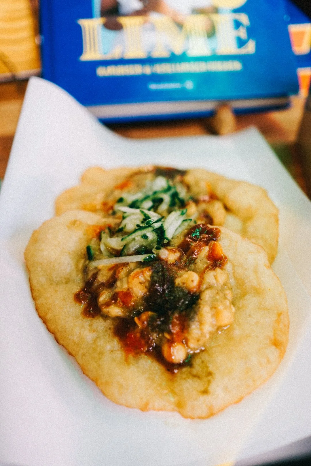 Close-up of a Chinese taco with fried dough, topped with vegetables, chicken, and sauce, on a white paper plate with a box of Lemon cookies in the background.