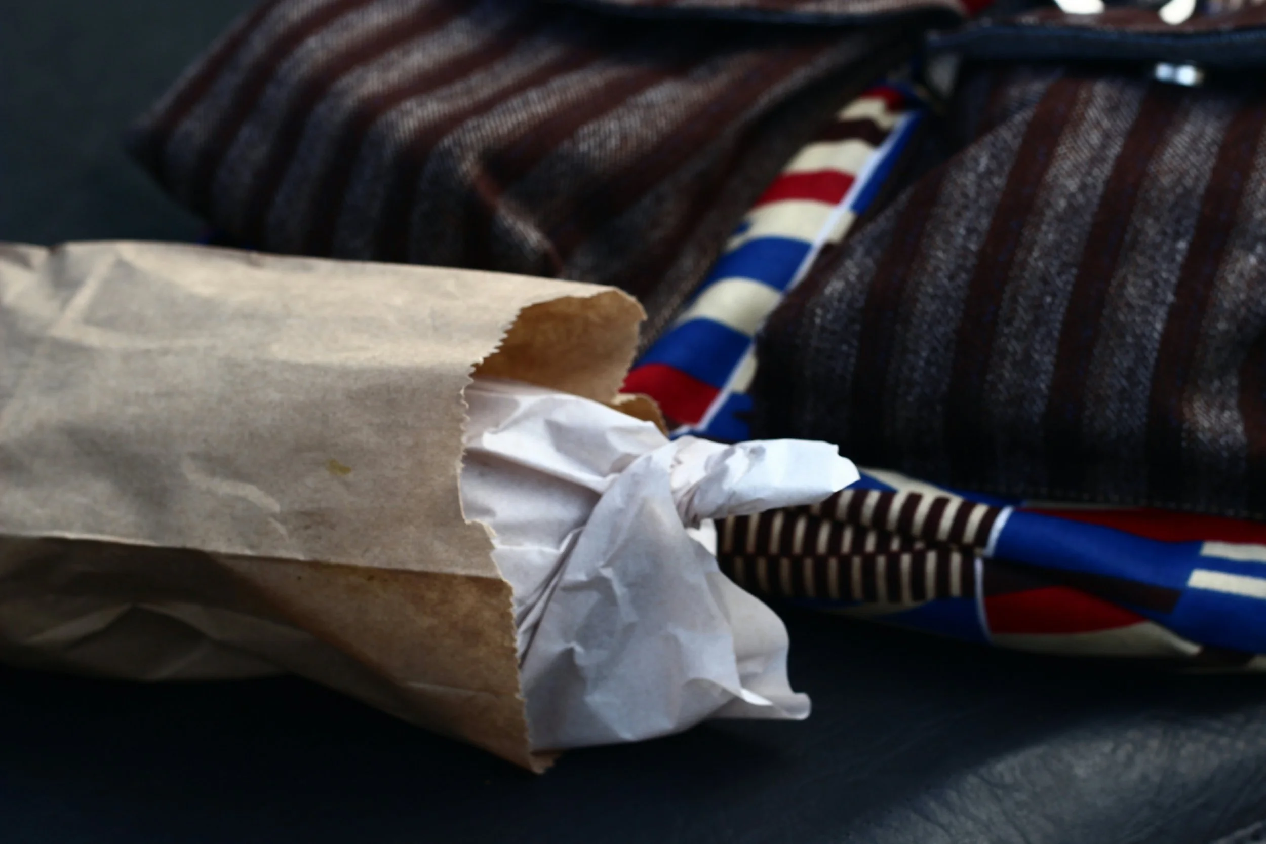 A Trini Doubles wrapped in paper placed on a dark surface, next to a striped a backpack with African wax print.