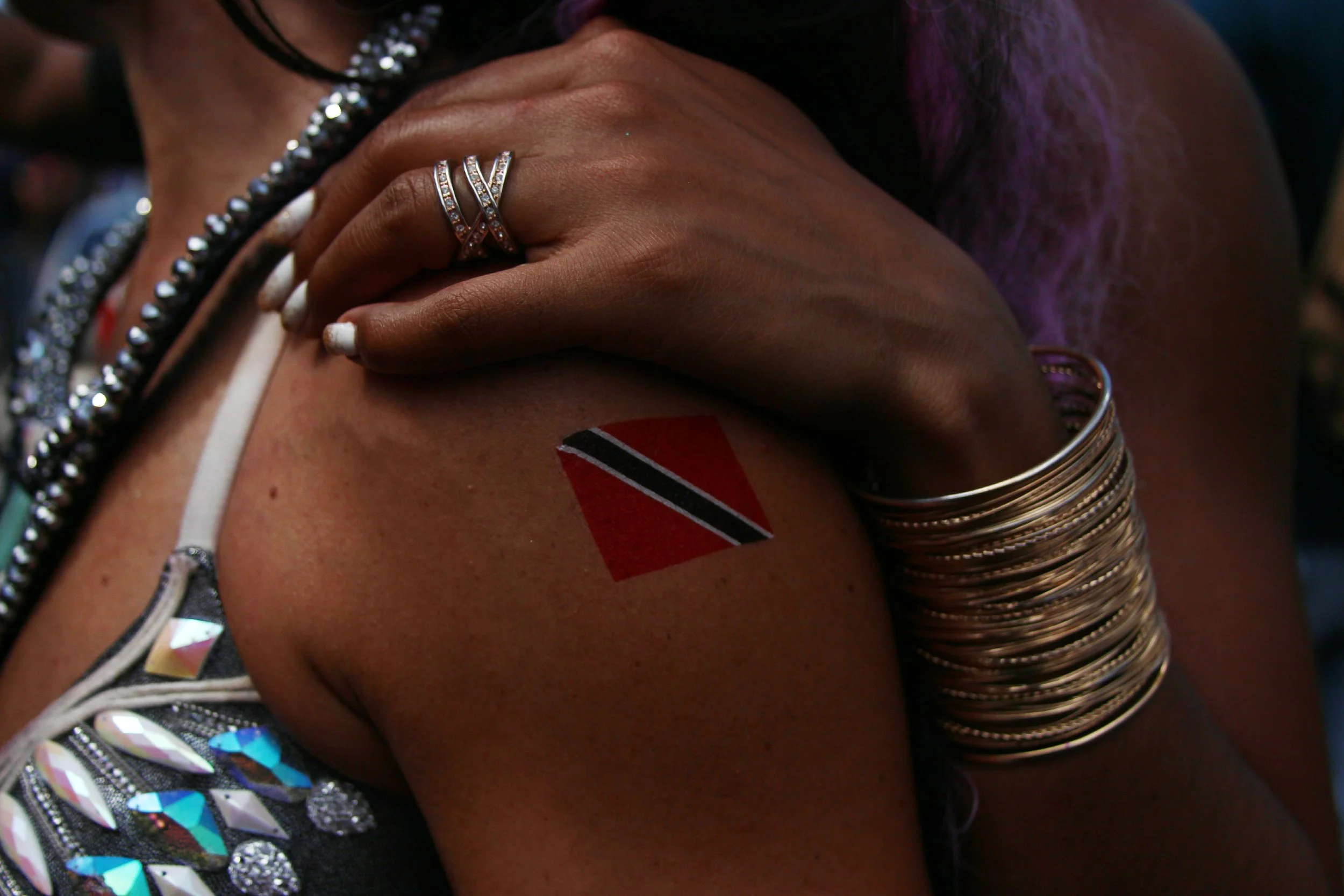 A woman with dark skin displays a temporary tattoo of the Trinidad and Tobago flag on her upper arm. She wears several gold bangles on her wrist, a ring on her finger, and has white nail polish.
