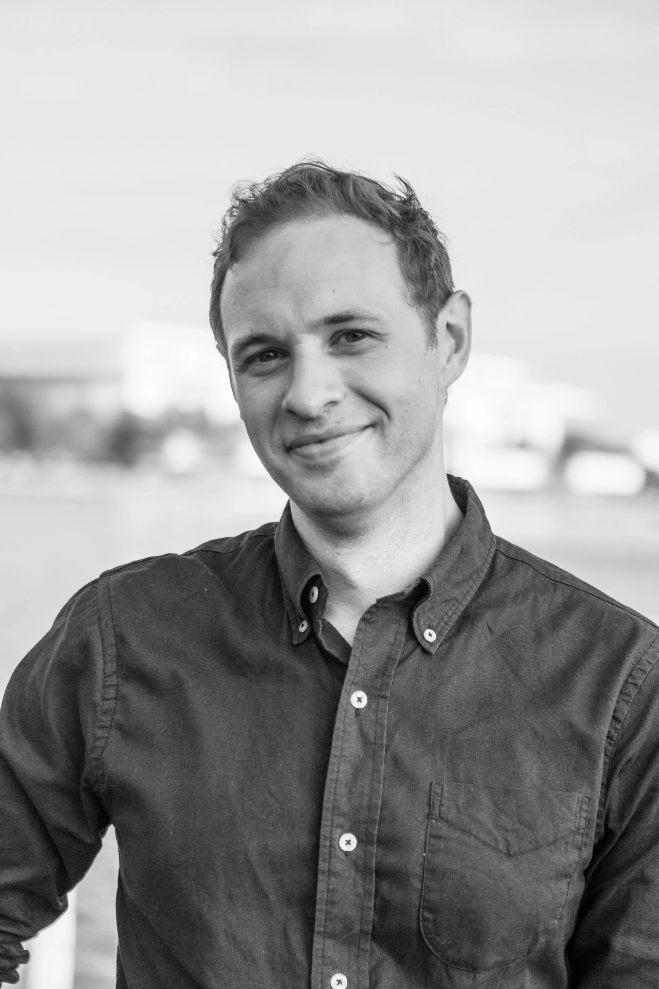 Black and white photo of a young man with short, wavy hair smiling outdoors, wearing a button-up shirt.