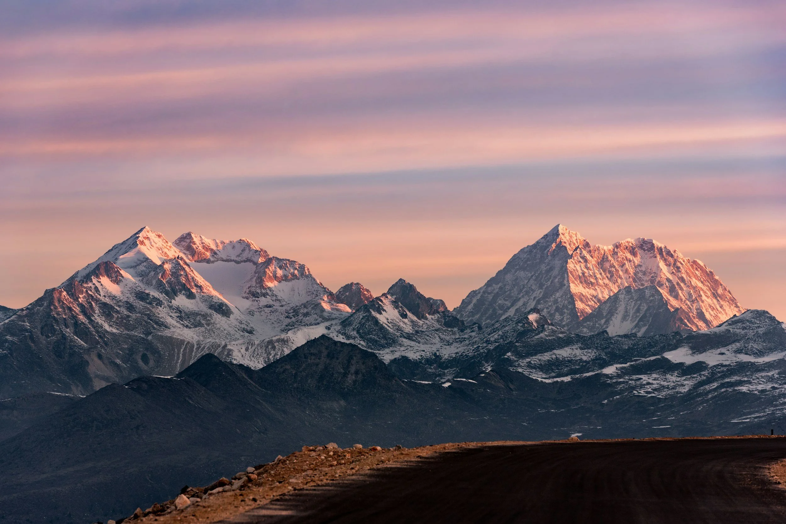 Snow-capped mountains at sunset with a colorful sky and a dark foreground road.