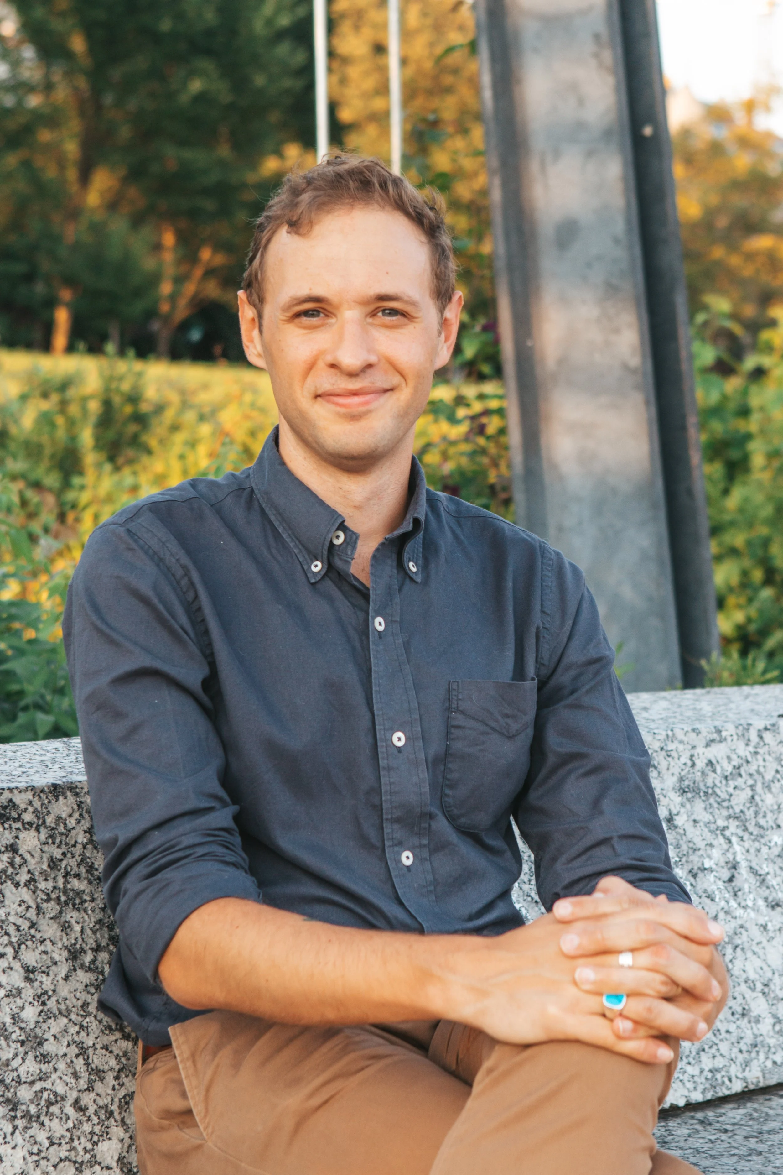 A young man with short light brown hair and blue eyes, smiling while sitting outdoors on a stone bench. He is wearing a dark blue button-up shirt and tan pants, with his hands clasped in front of him. The background features greenery and autumn-colored trees.
