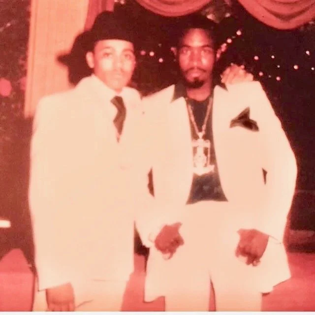 Two men in white suits standing side by side in front of a decorated curtain, one with curly hair and the other with braids, at a formal event.