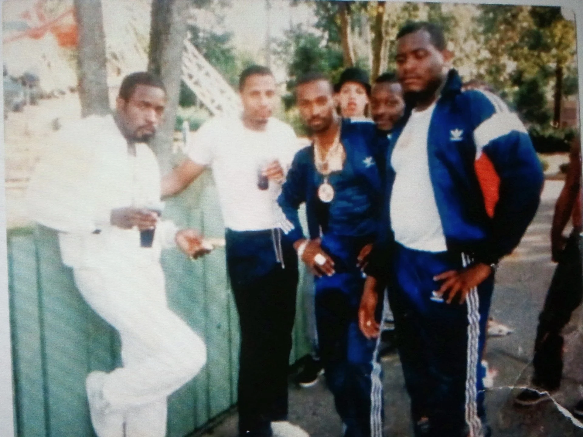 Five men are gathered outdoors near a green wooden fence, with trees in the background. One man is dressed in all white, holding a drink and standing with one foot on the fence. The other four men are wearing matching blue Adidas tracksuits and have