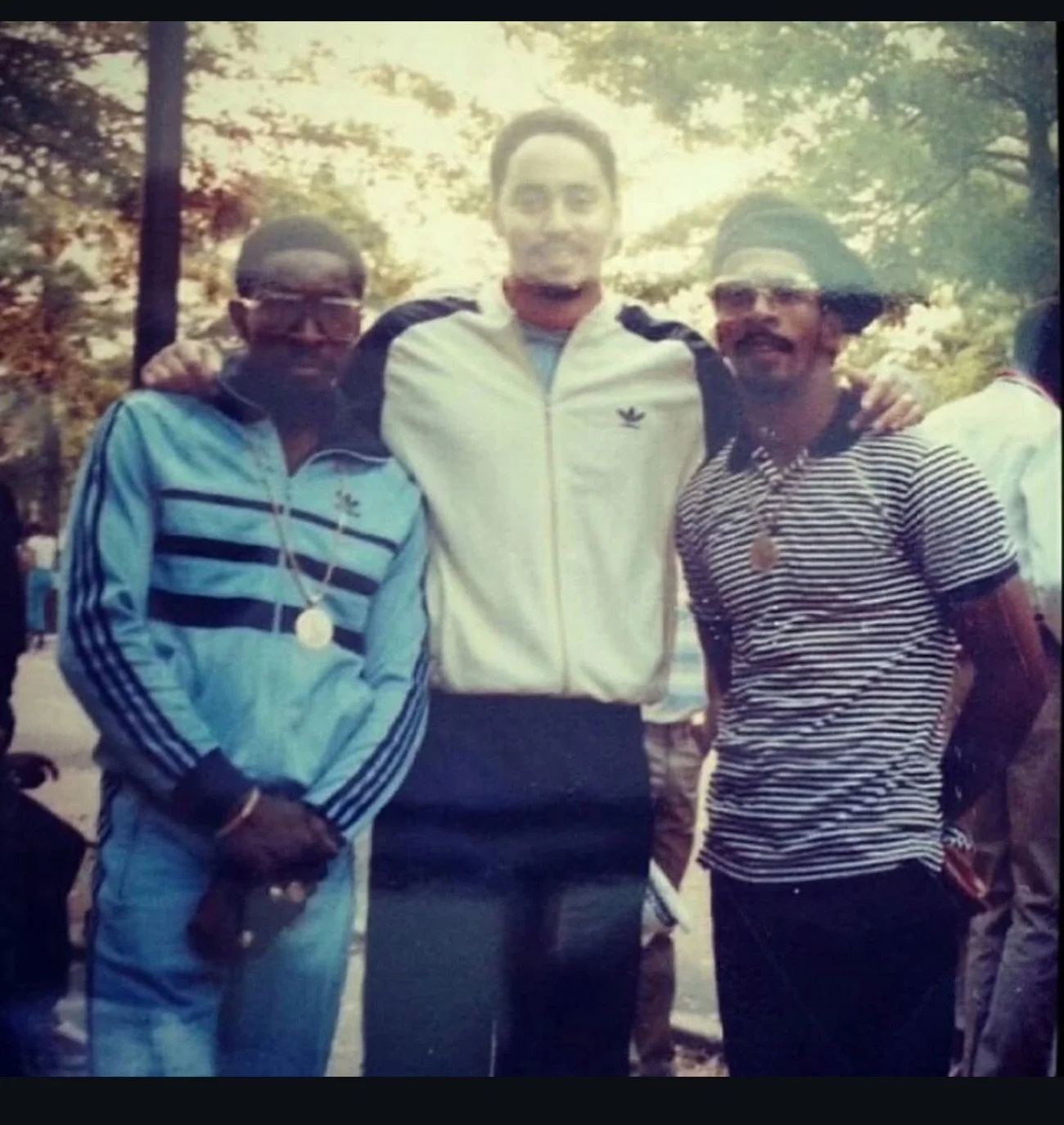 Three men standing outdoors surrounded by trees, posing for a photo. The man on the left is wearing glasses, a light blue and black striped jacket, and holding a gun. The man in the middle is taller, wearing a white jacket with black and blue accents
