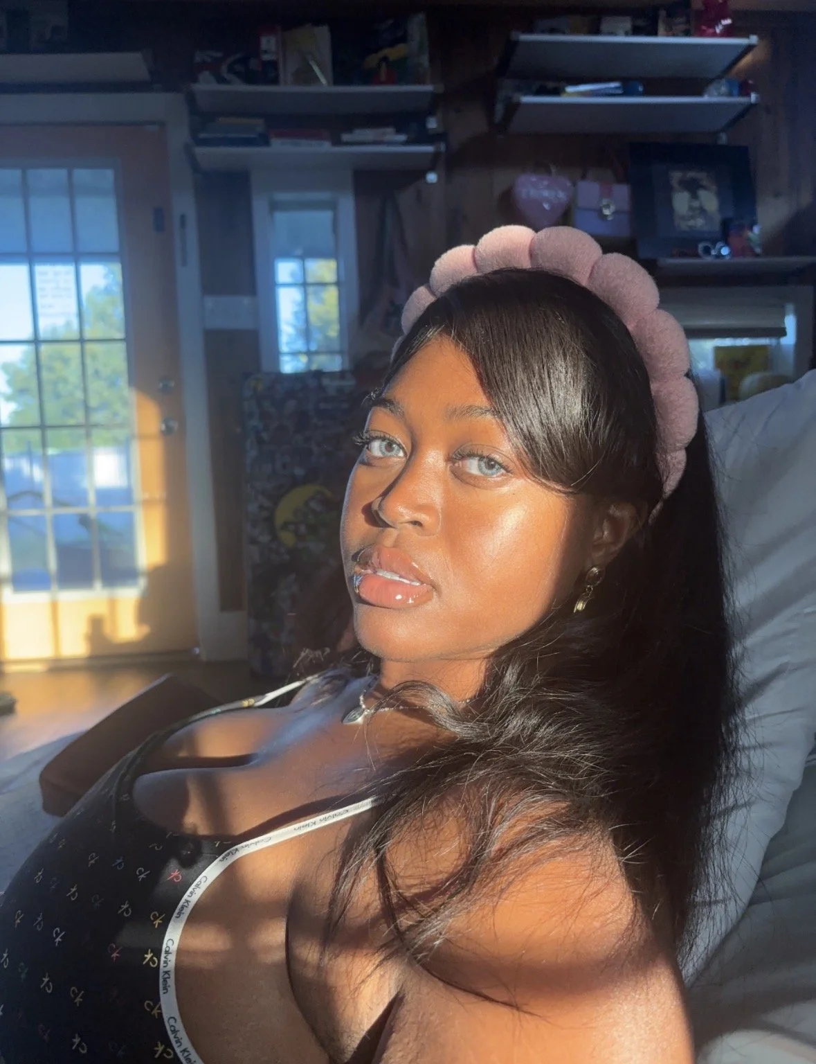 Kenya Brown taking an Instagram photo wearing a pink fluffy headband used for skincare from Amazon and a thrifted Calvin Klein pajama tank top. In her artist loft with Easter orange, green and yellow walls,