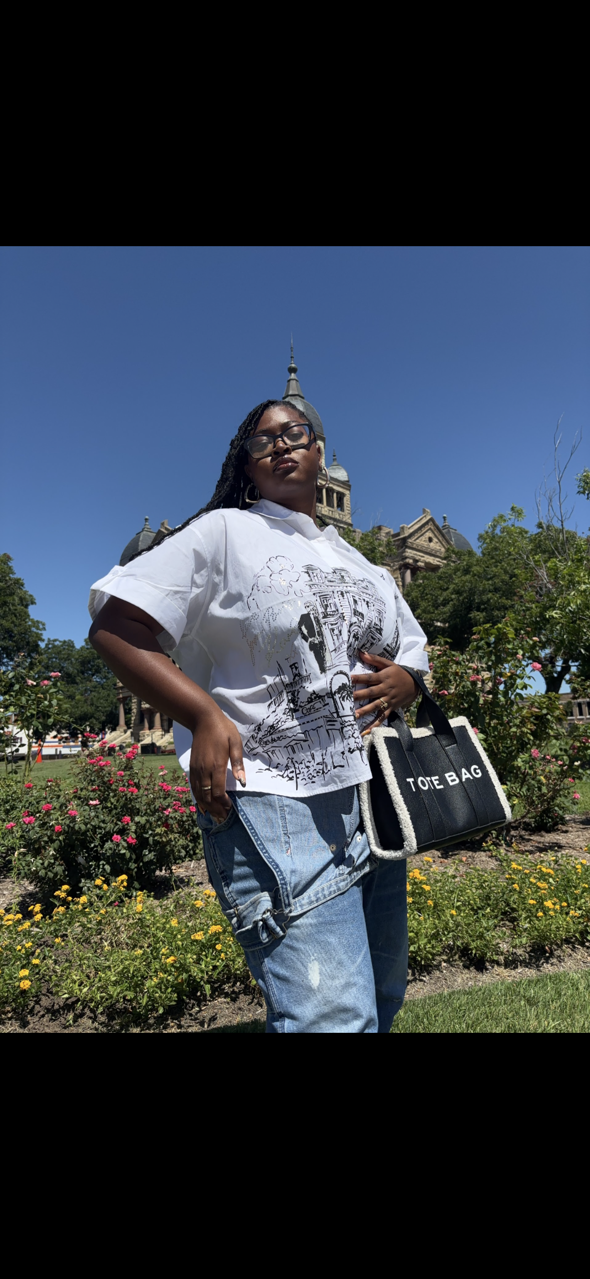 A woman wearing sunglasses, a white graphic shirt, and ripped jeans stands in front of a historic building with a clock tower, holding a black tote bag with white text, amidst a garden with pink and yellow flowers and green trees under a clear blue s