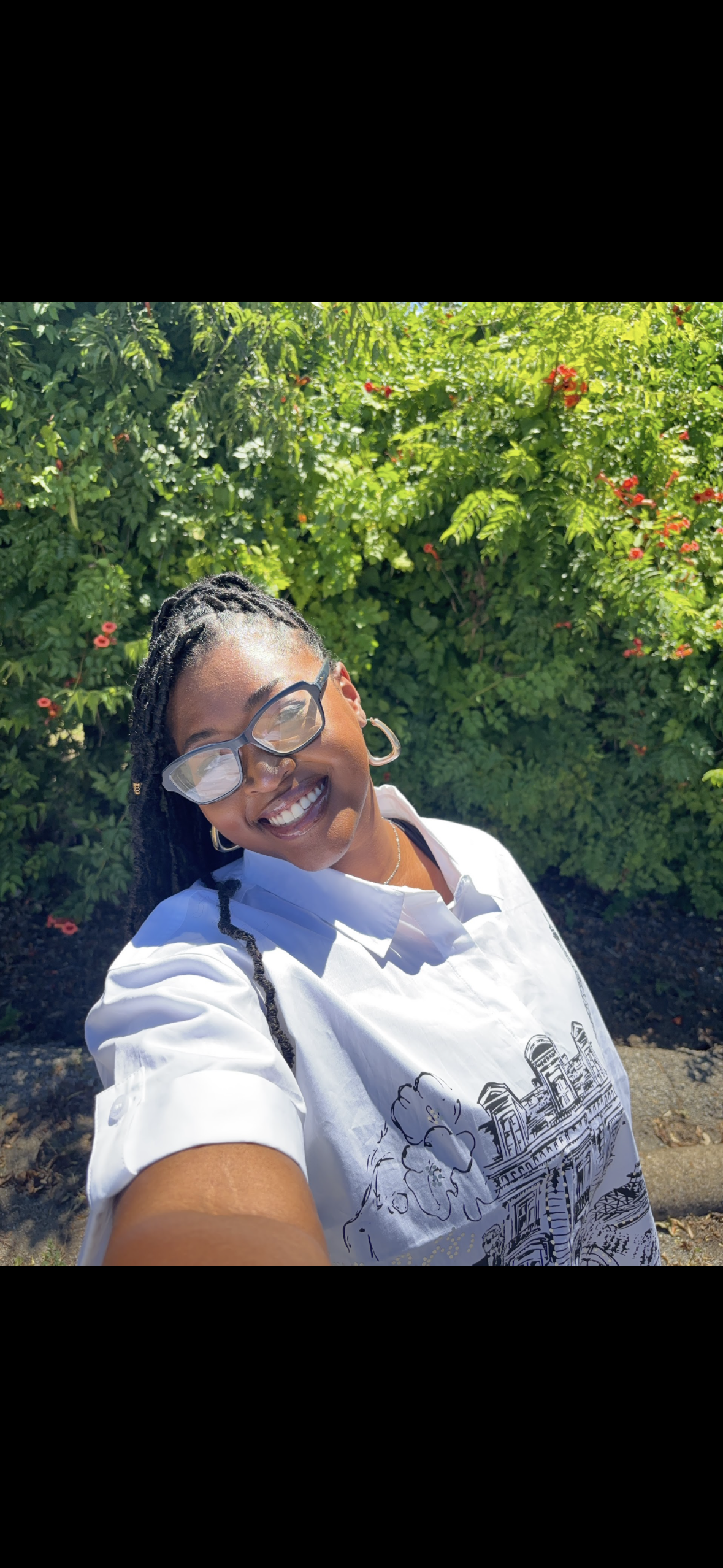 Kenya Brown taking a selfie outdoors, smiling, wearing glasses and a white shirt with a black line drawing of buildings and flowers, standing in front of green bushes with small red flowers.