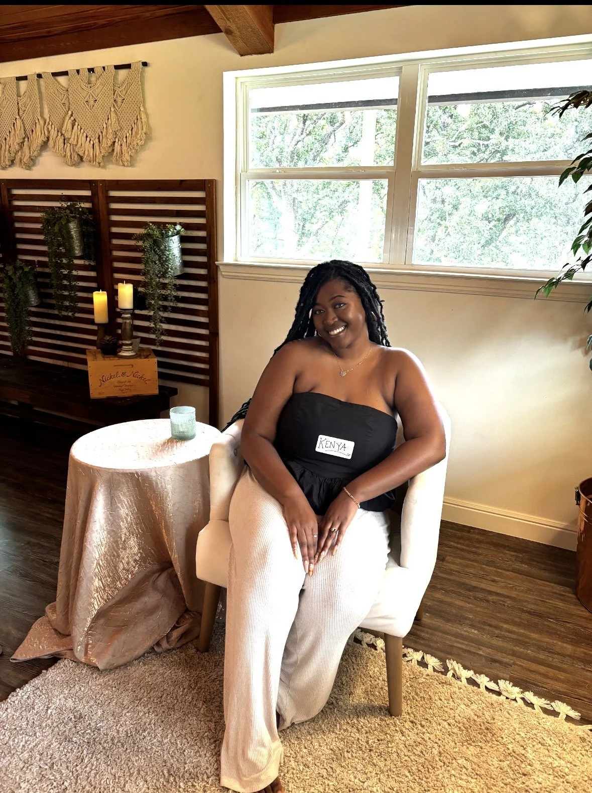 A woman sitting on a white chair indoors next to a small round table covered with a shiny pink cloth, with a candle holder and candles on it. She is smiling, wearing a black strapless top with a name tag that reads 'Kenya' and light-colored pants. Th