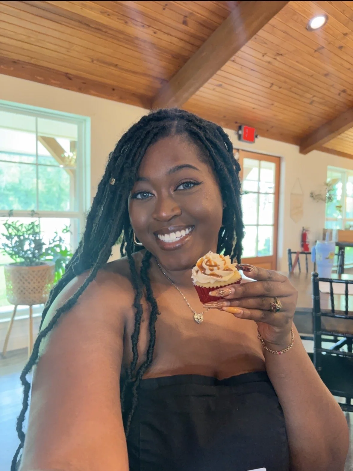 A woman with long dreadlocks smiling while holding a cupcake topped with orange and white frosting in a well-lit room with wooden ceiling beams and large windows.