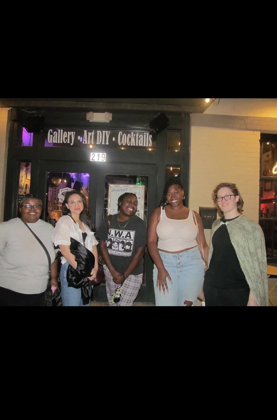 Group of five women standing outside a gallery, art DIY, and cocktails bar at night, smiling.