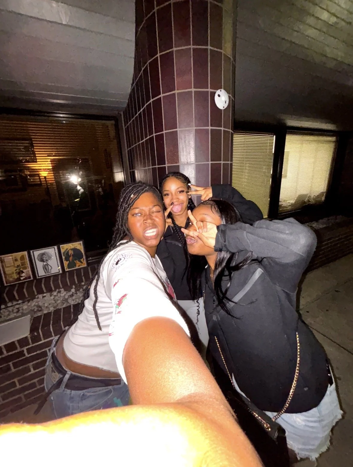 Three women posing for a selfie outside a building at night, making playful facial expressions and gestures.