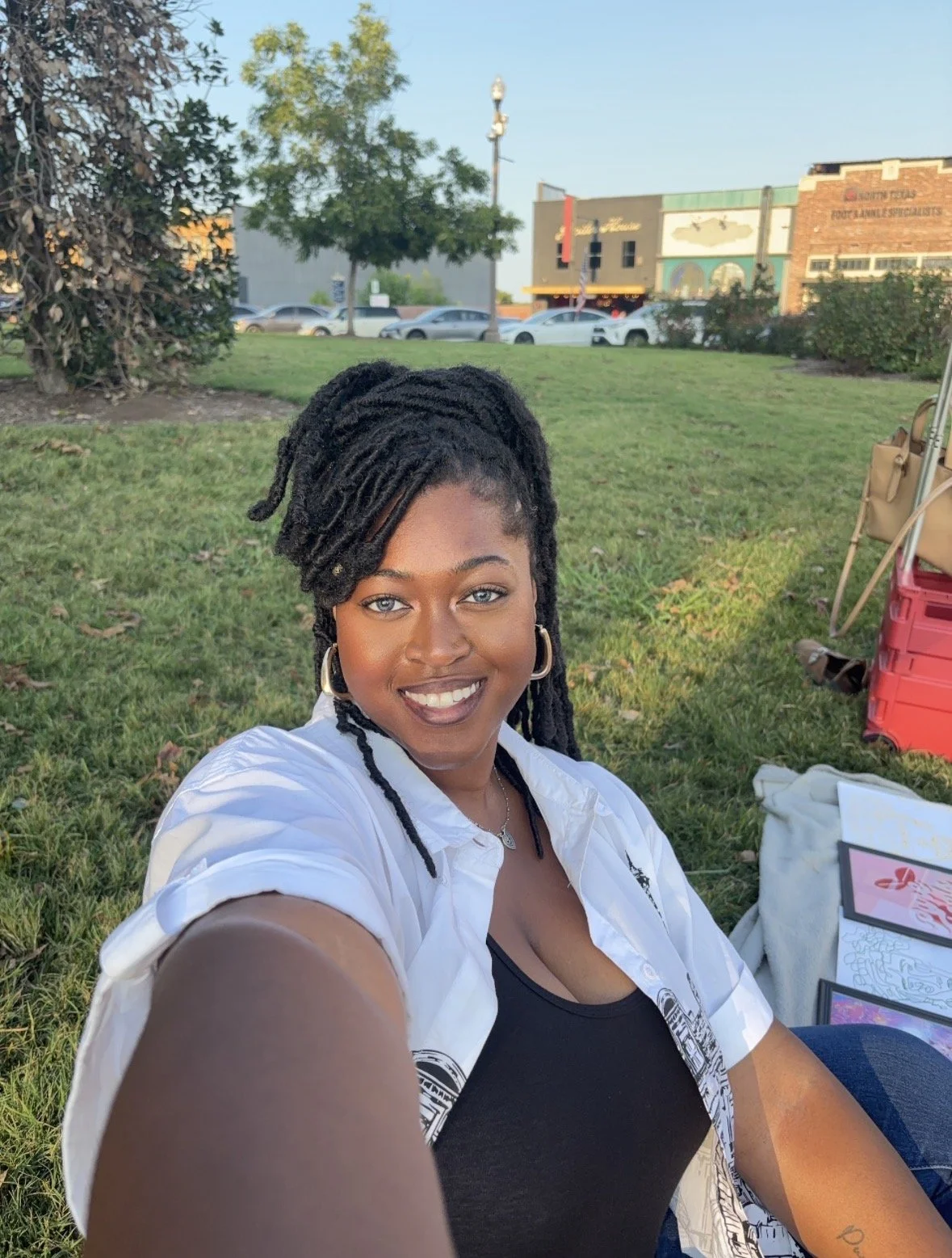 A woman with dark skin and dreadlocks taking a selfie outdoors, sitting on grass with a background of trees, buildings, cars, and a clear sky.
