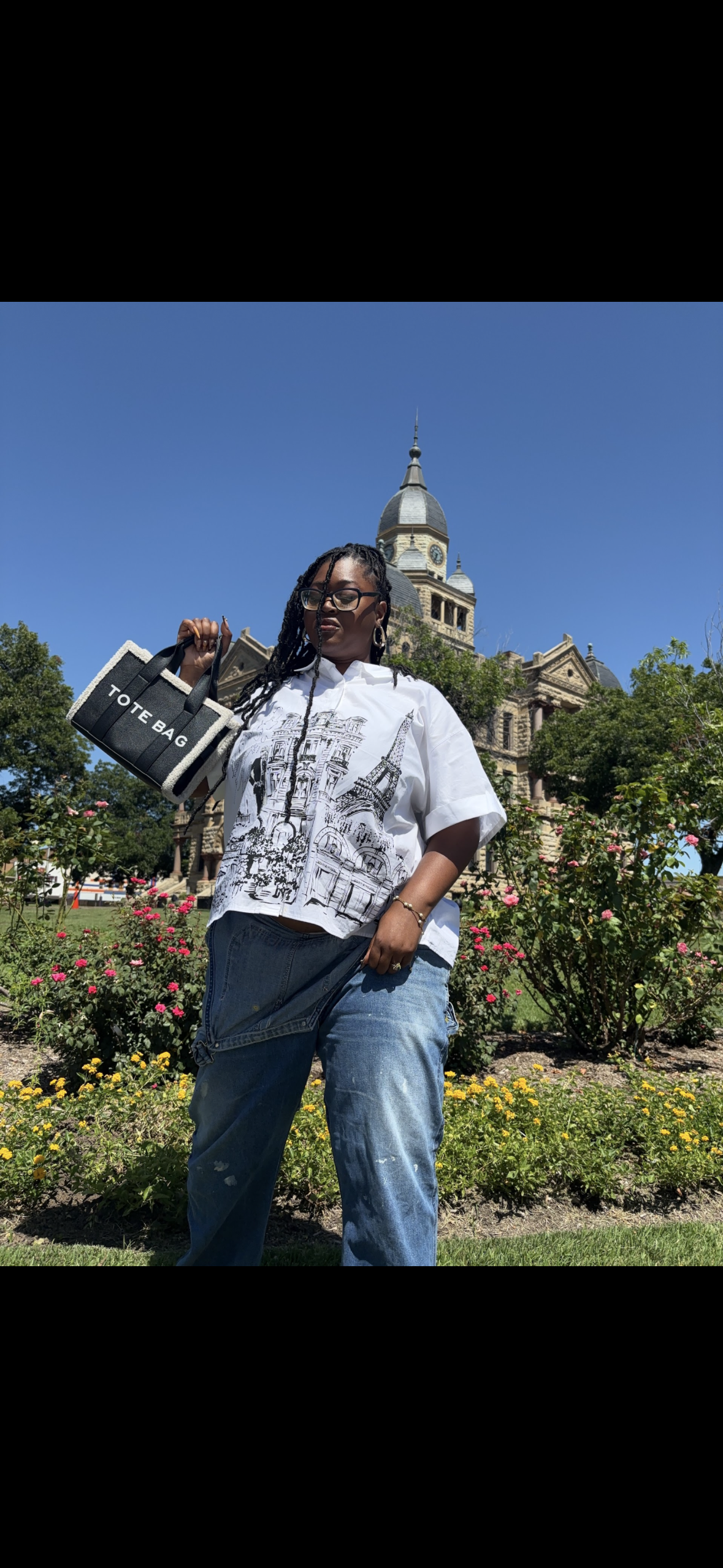 A woman standing outdoors in front of a historic building with a clock tower, wearing a white graphic shirt, ripped jeans, glasses, and holding a tote bag.
