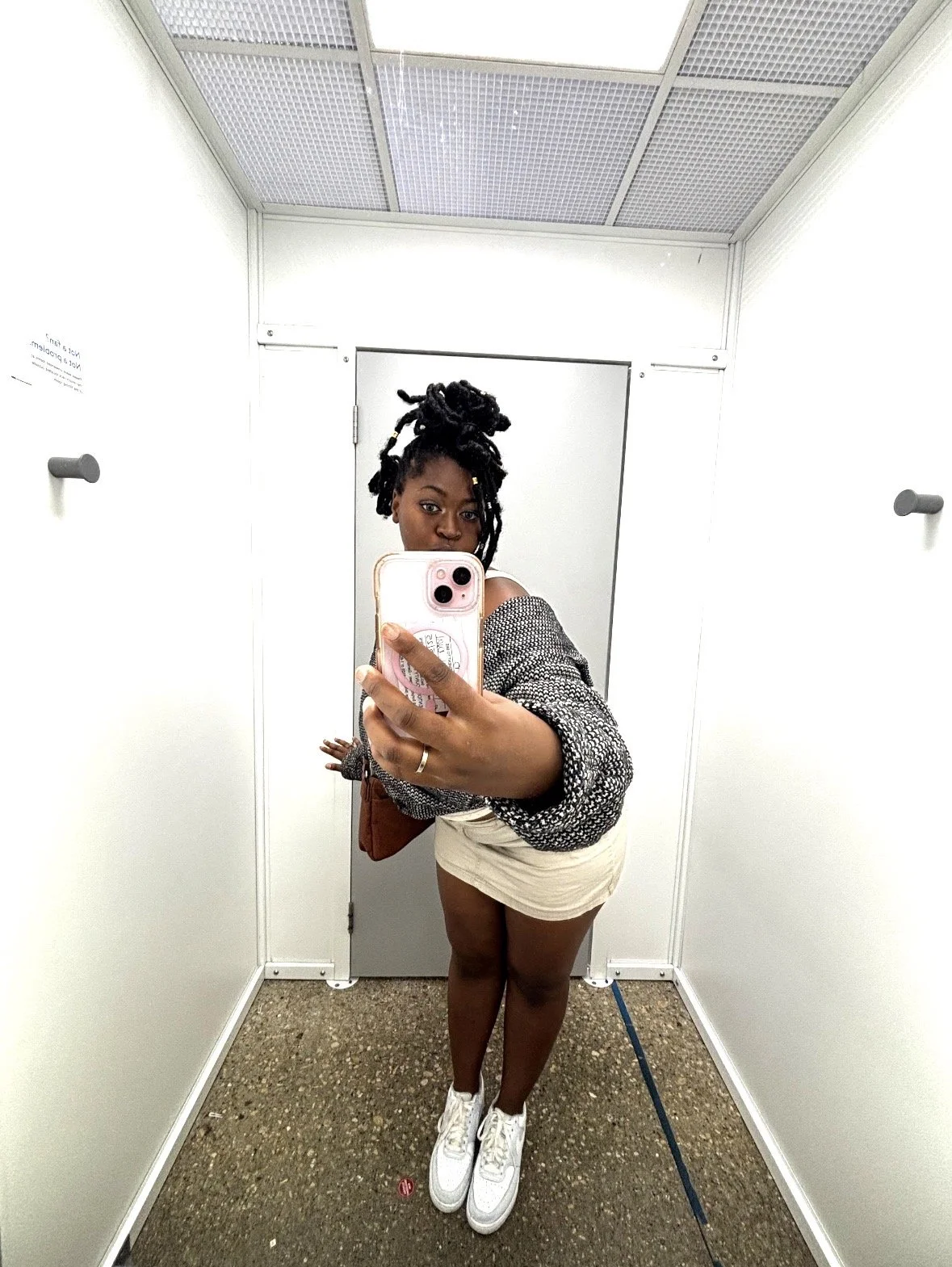 Kenya Brown modeling Walmart fashion with dreadlocks taking a selfie in a fitting room mirror wearing a black and white patterned jacket, beige shorts, and white Nike sneakers.