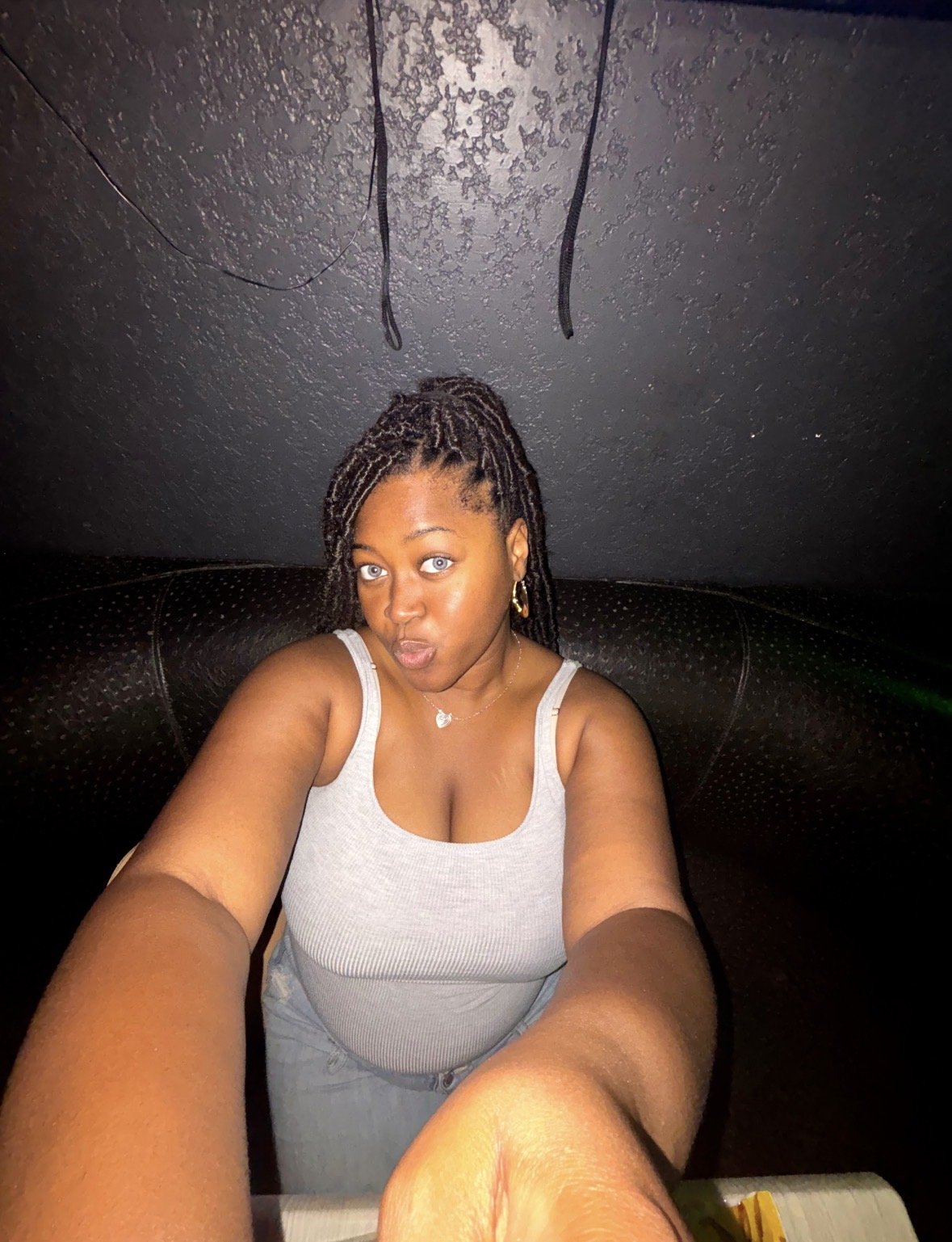 A woman with braids and blue eyes taking a selfie in a dimly lit room, wearing a gray tank top and gold jewelry, seated on a black leather couch.