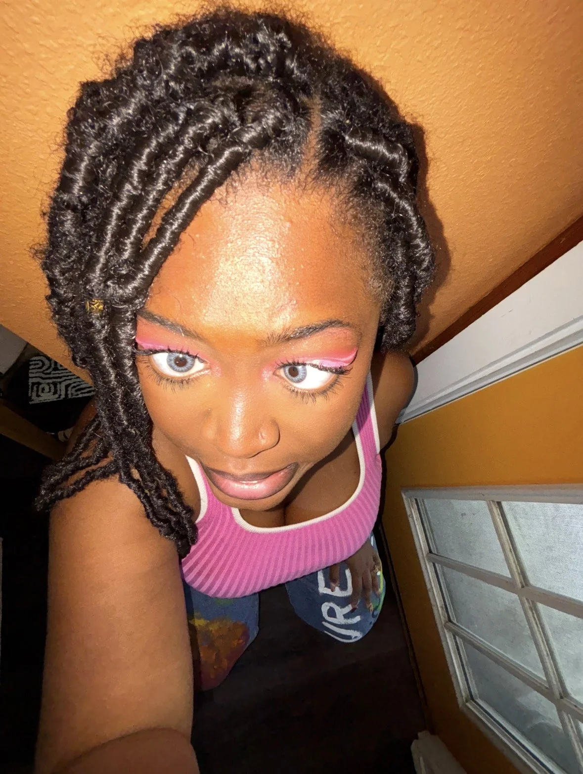 A woman with styled hair, wearing pink eyeshadow and a pink and white striped tank top, looking up at the camera from an overhead angle inside a room with a window and orange walls.