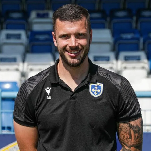 A sports physio in a black sports polo shirt with a sports team logo on the chest, standing in front of blue and white stadium seats.