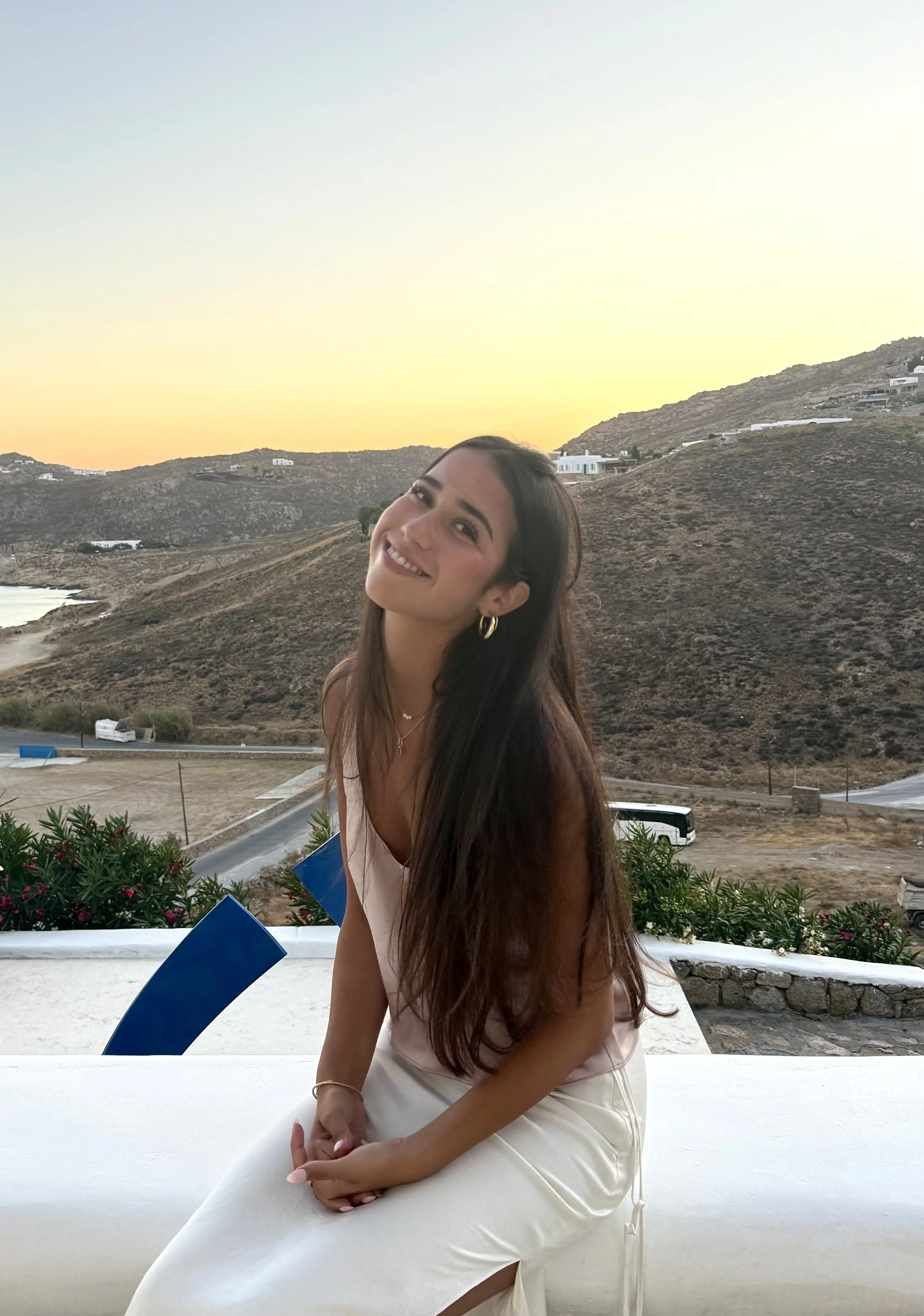 A woman with long dark hair smiling, sitting outdoors on a white ledge, with a hilly landscape and white buildings in the background during sunset.