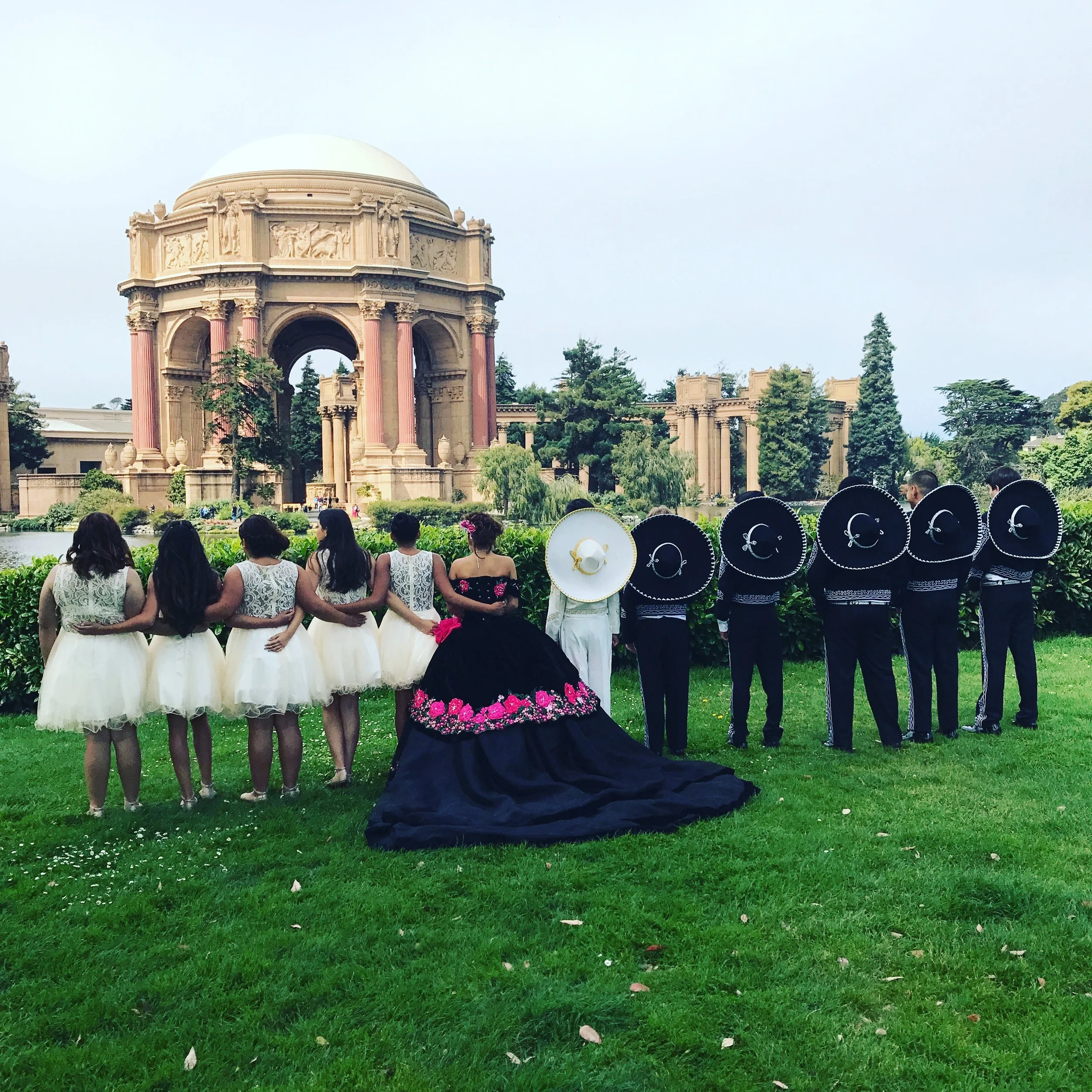 Group of people, including women in white tutus and men in black outfits with traditional Mexican sombreros, standing on grass in front of the Palace of Fine Arts in Mexico City, with a large domed building in the background.