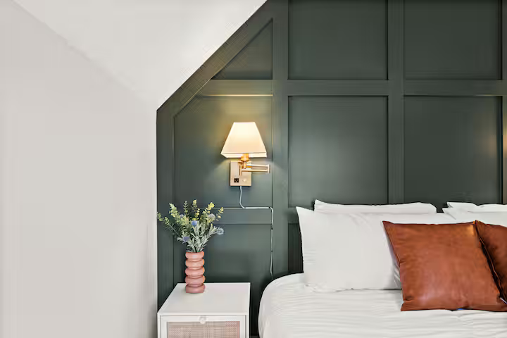 A bedroom with a white bedside table holding a pink vase with greenery, a wall-mounted lamp, and a bed with white linens and brown pillows.